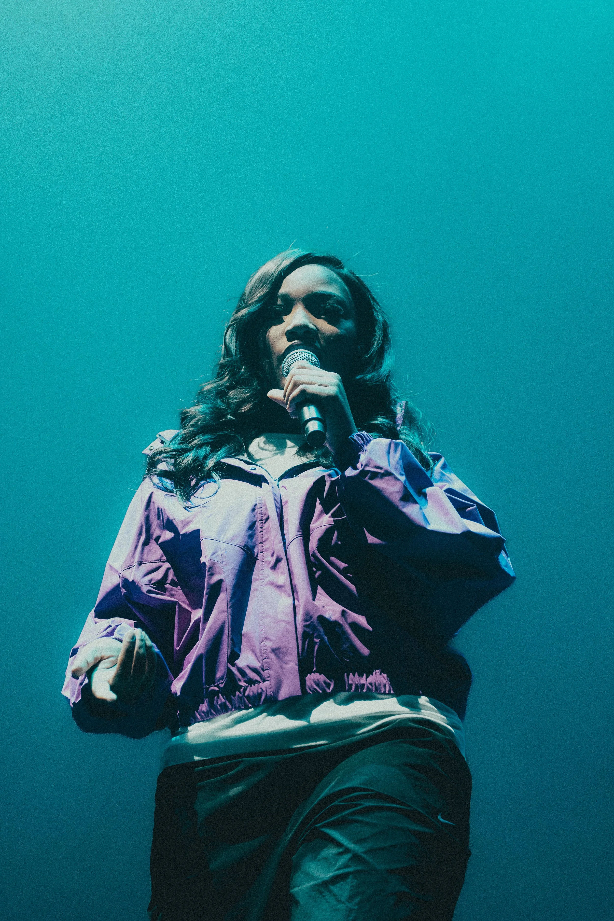 Femme qui chante dans un micro, portant une veste violette, sur scène avec un éclairage vert