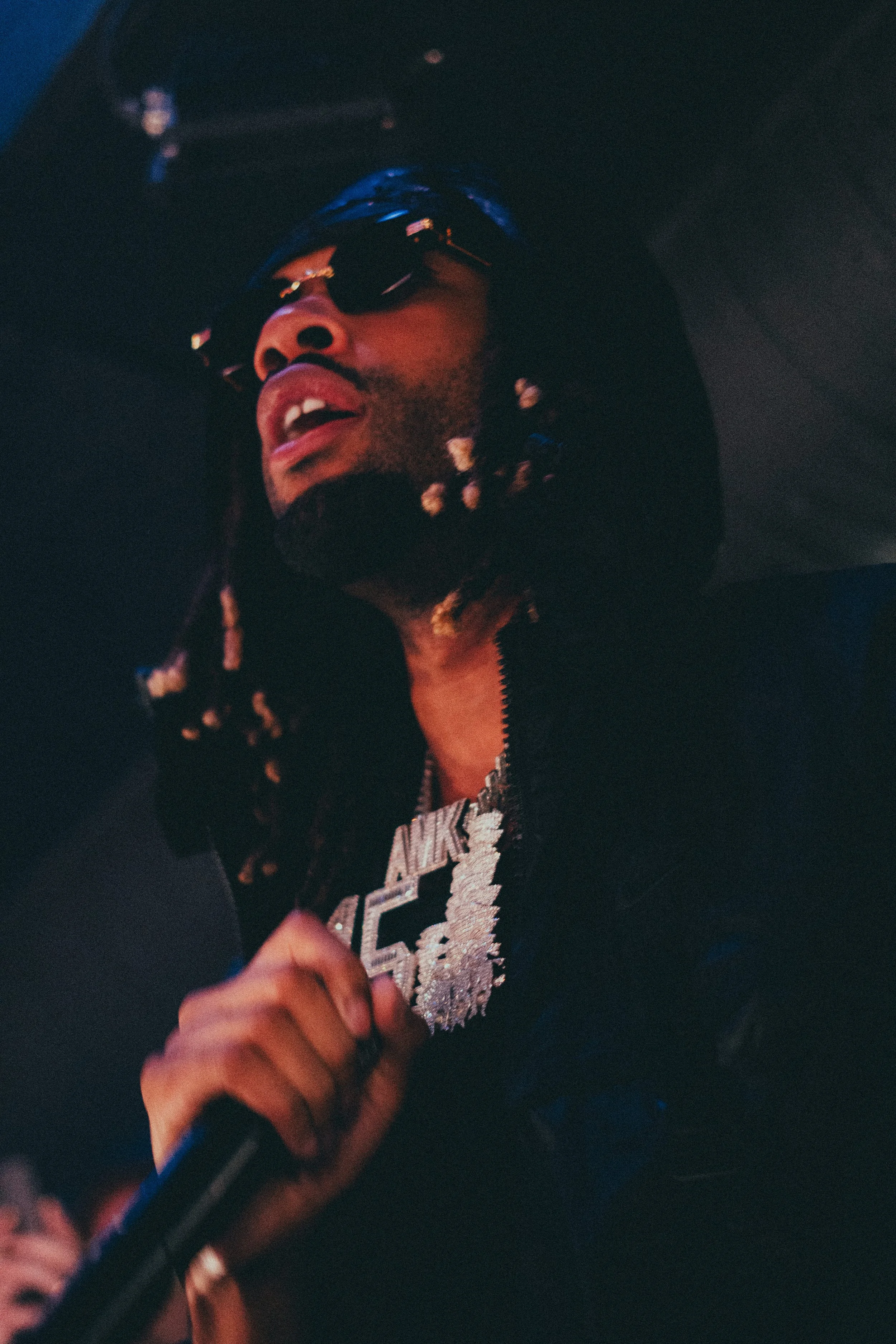 Un homme portant des lunettes de soleil, un bonnet, un collier en diamants avec la inscription 'ANK' et un sweat à capuche noir, tenant un micro dans une main.