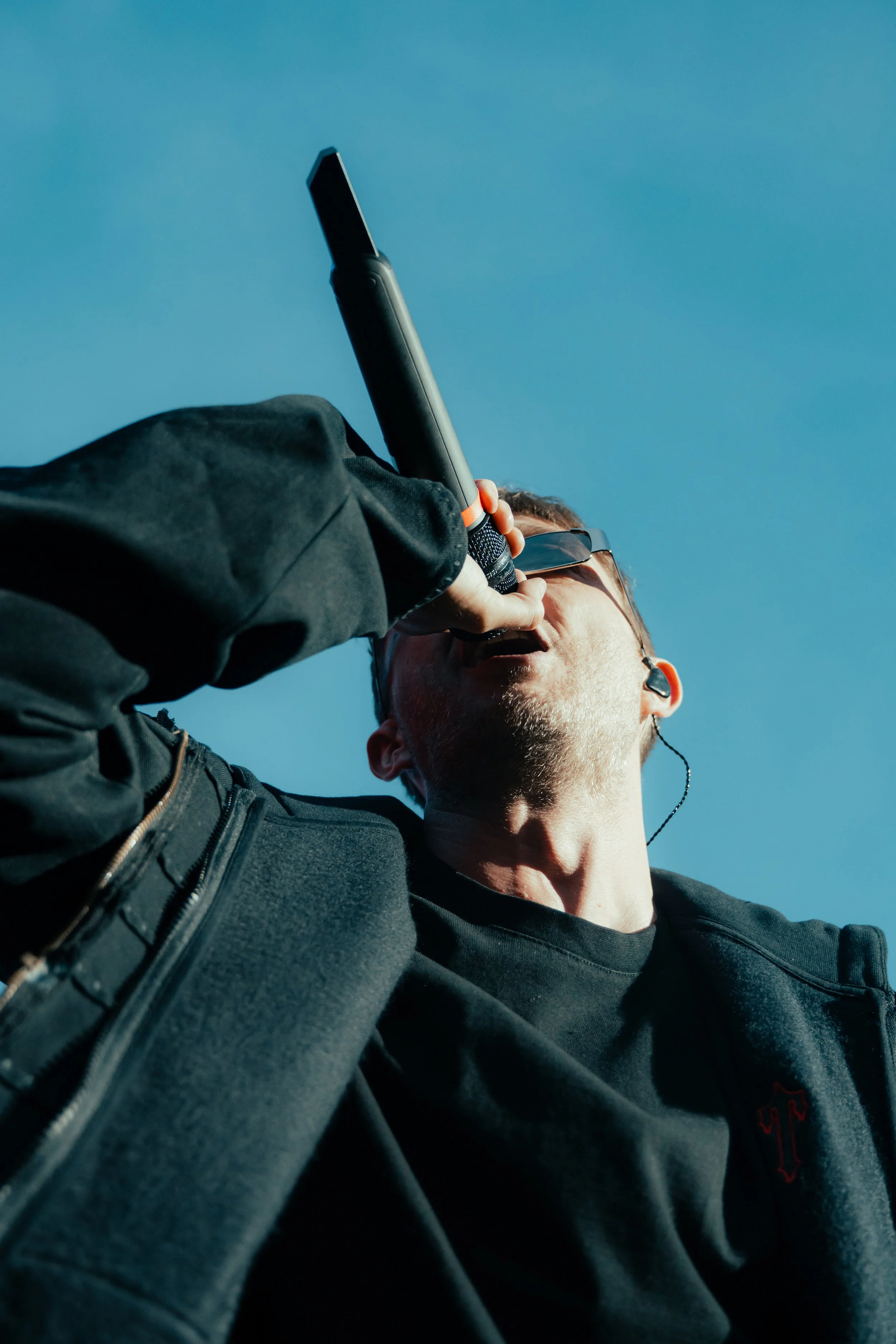 Un homme portant des lunettes de soleil et des écouteurs, vêtu d'une veste noire, parlant dans un microphone avec un ciel bleu en arrière-plan.