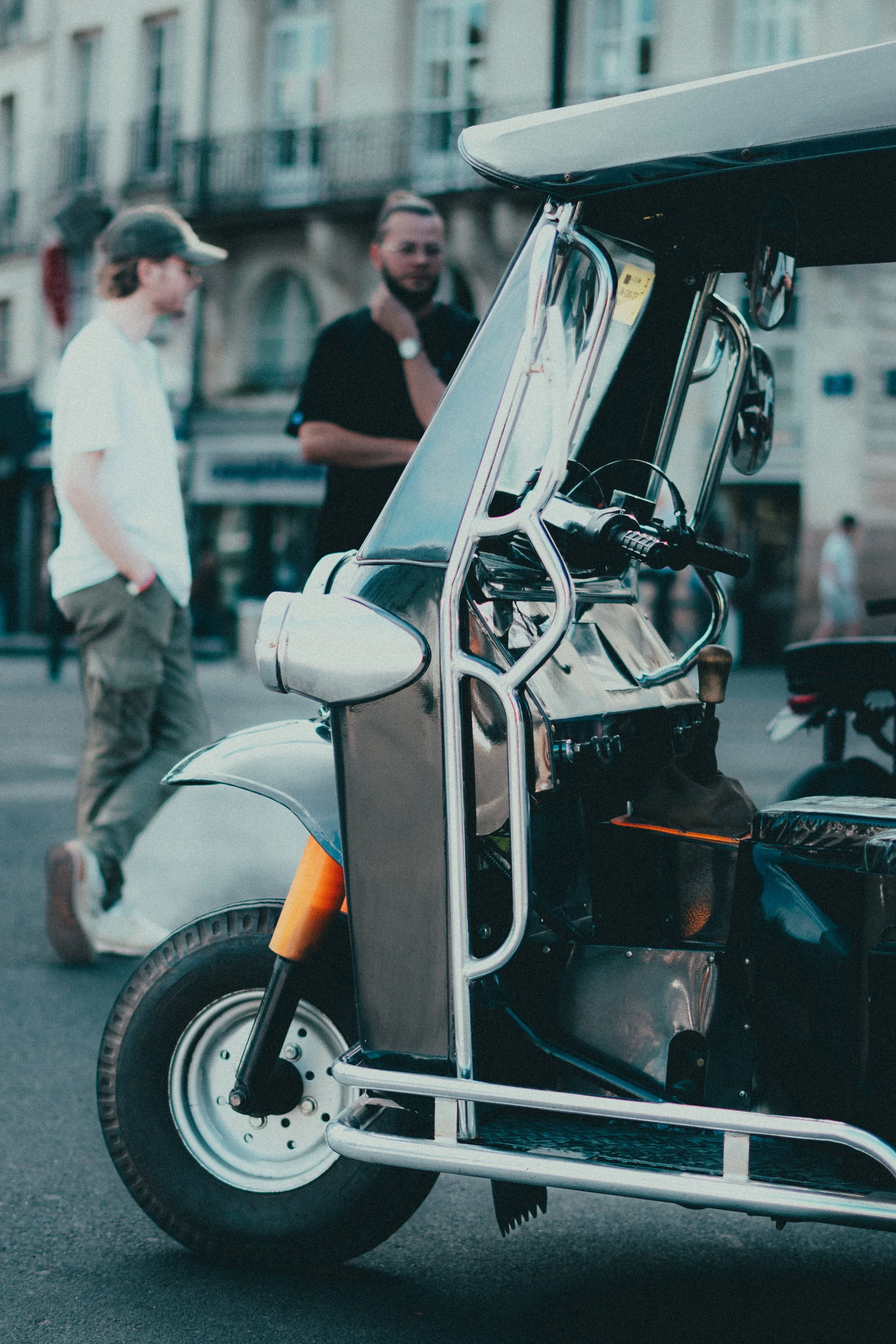 Un triporteur électrique stationné sur la rue, avec deux hommes en arrière-plan, l'un portant une casquette blanche et l'autre avec une barbe et une montre.