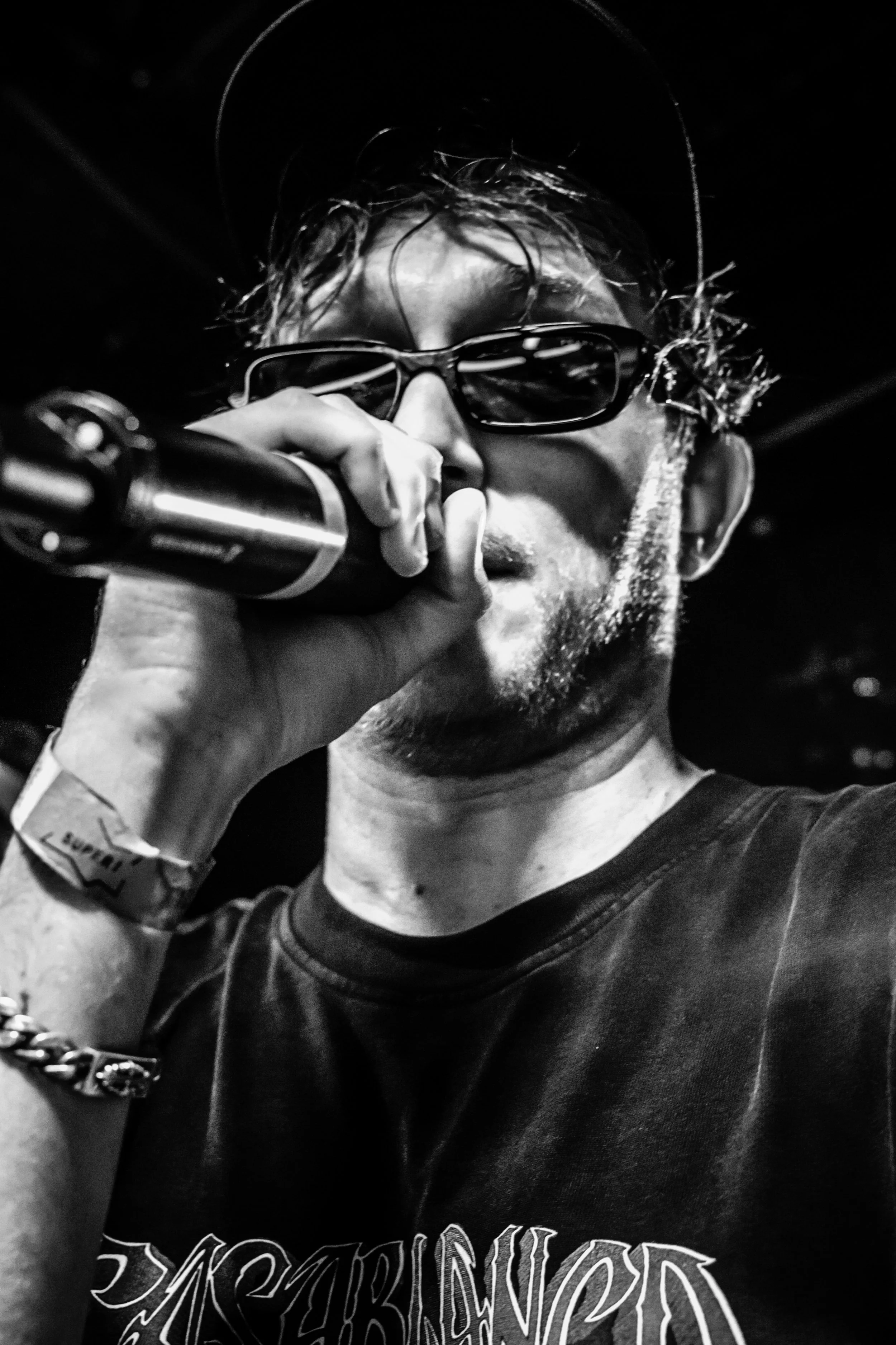 Un homme portant des lunettes de soleil, une casquette, et une chemise noire, tenant un microphone, en train de chanter ou parler sur scène dans une ambiance sombre.