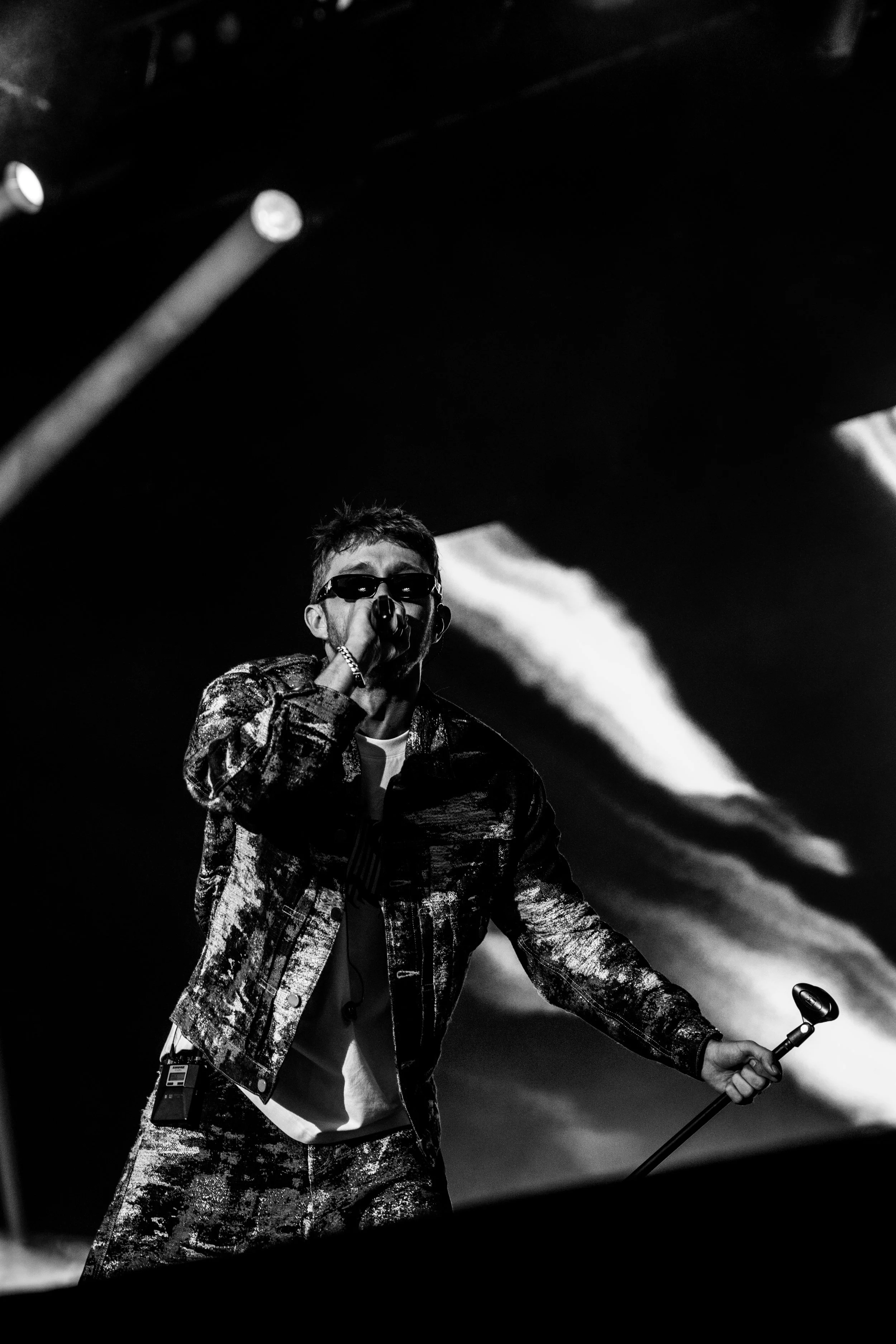 Un chanteur portant des lunettes de soleil et un collier, chantant ou parlant dans un microphone lors d'un concert, en noir et blanc.