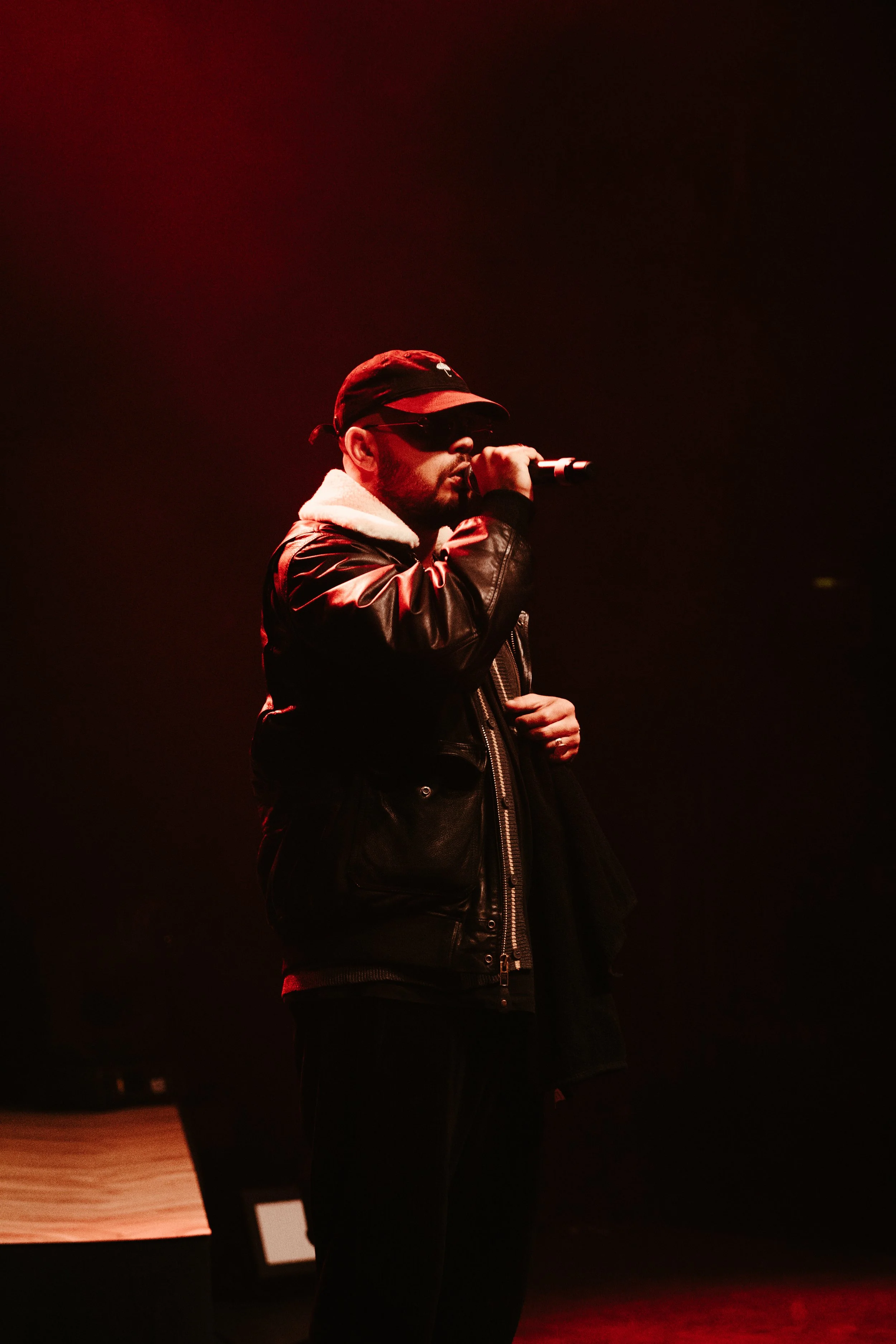 Un homme portant une veste en cuir, une casquette noire, des lunettes de soleil, tenant un micro sur scène avec un éclairage rougeâtre.