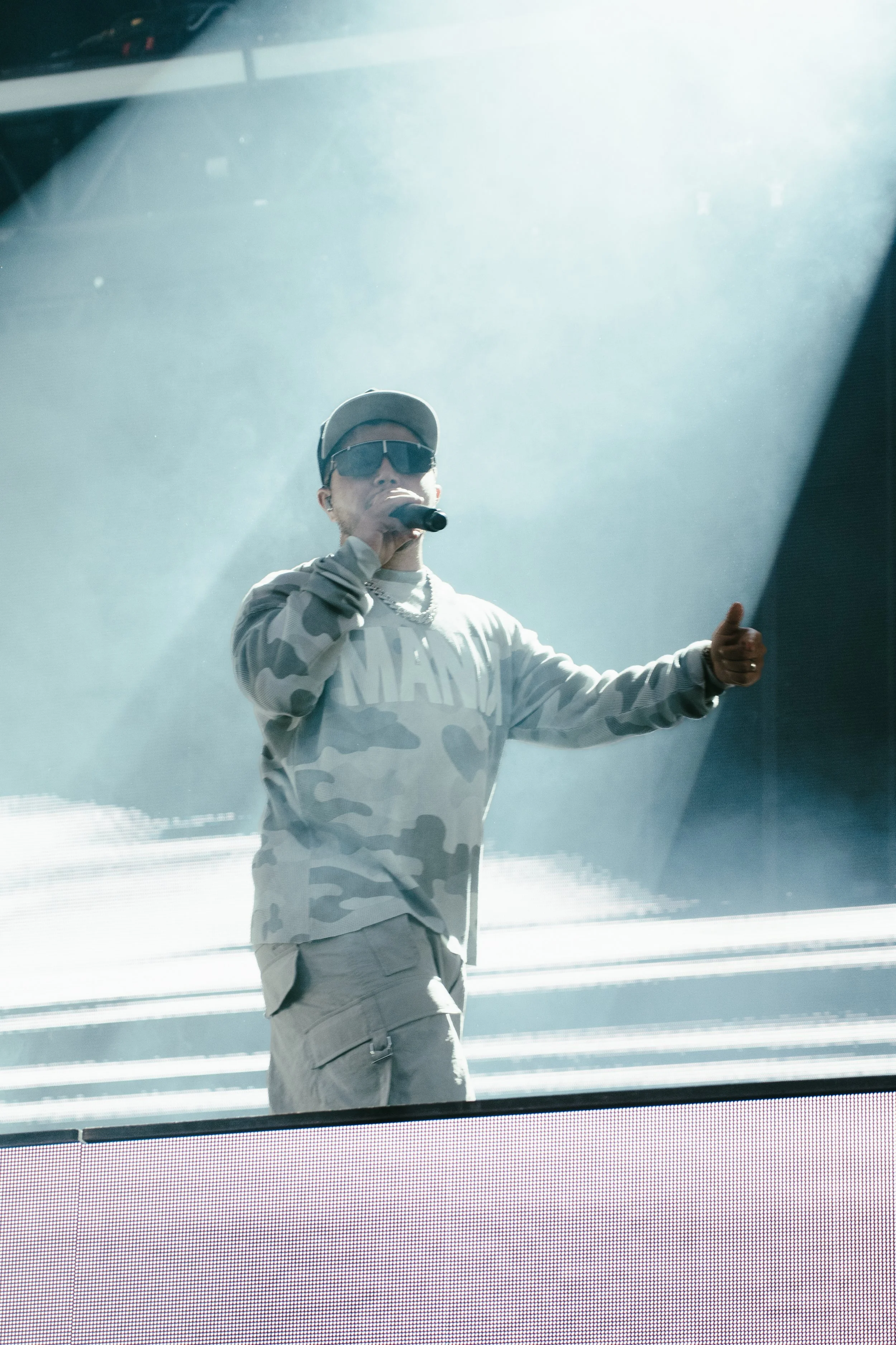 Un homme en tenues décontractées, portant une casquette et des lunettes de soleil, tient un microphone en scène, probablement lors d'un concert ou d'un événement en direct.