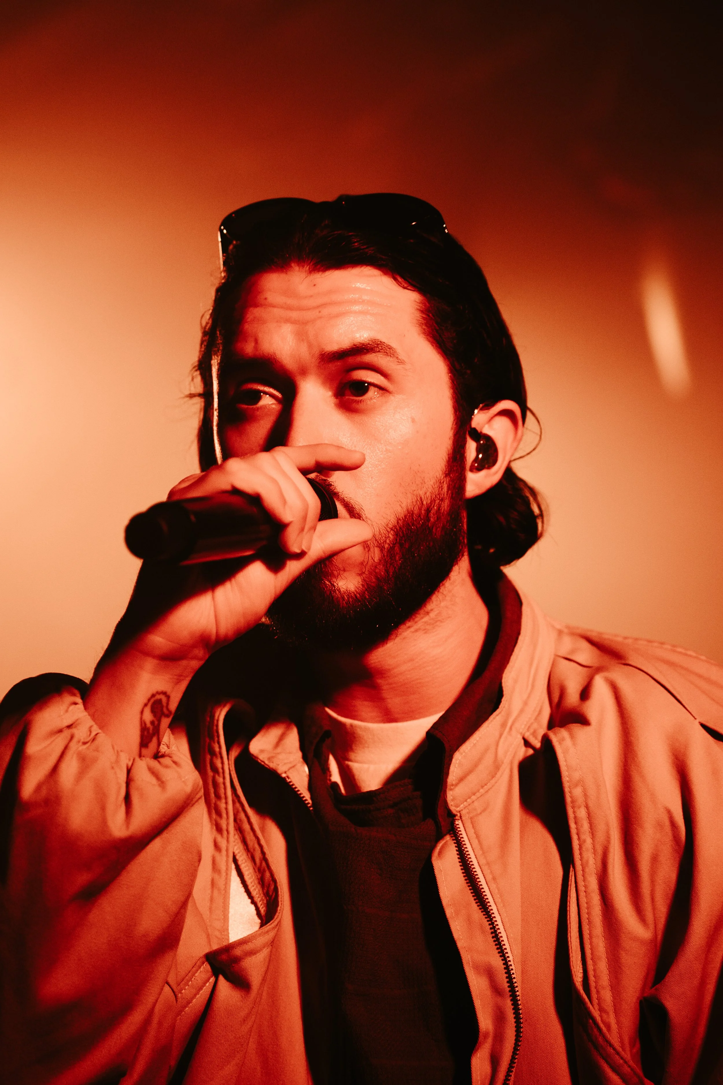 Un homme avec une barbe et des cheveux longs, portant des lunettes de soleil sur la tête, chante ou parle dans un microphone dans un éclairage rouge/orangé.