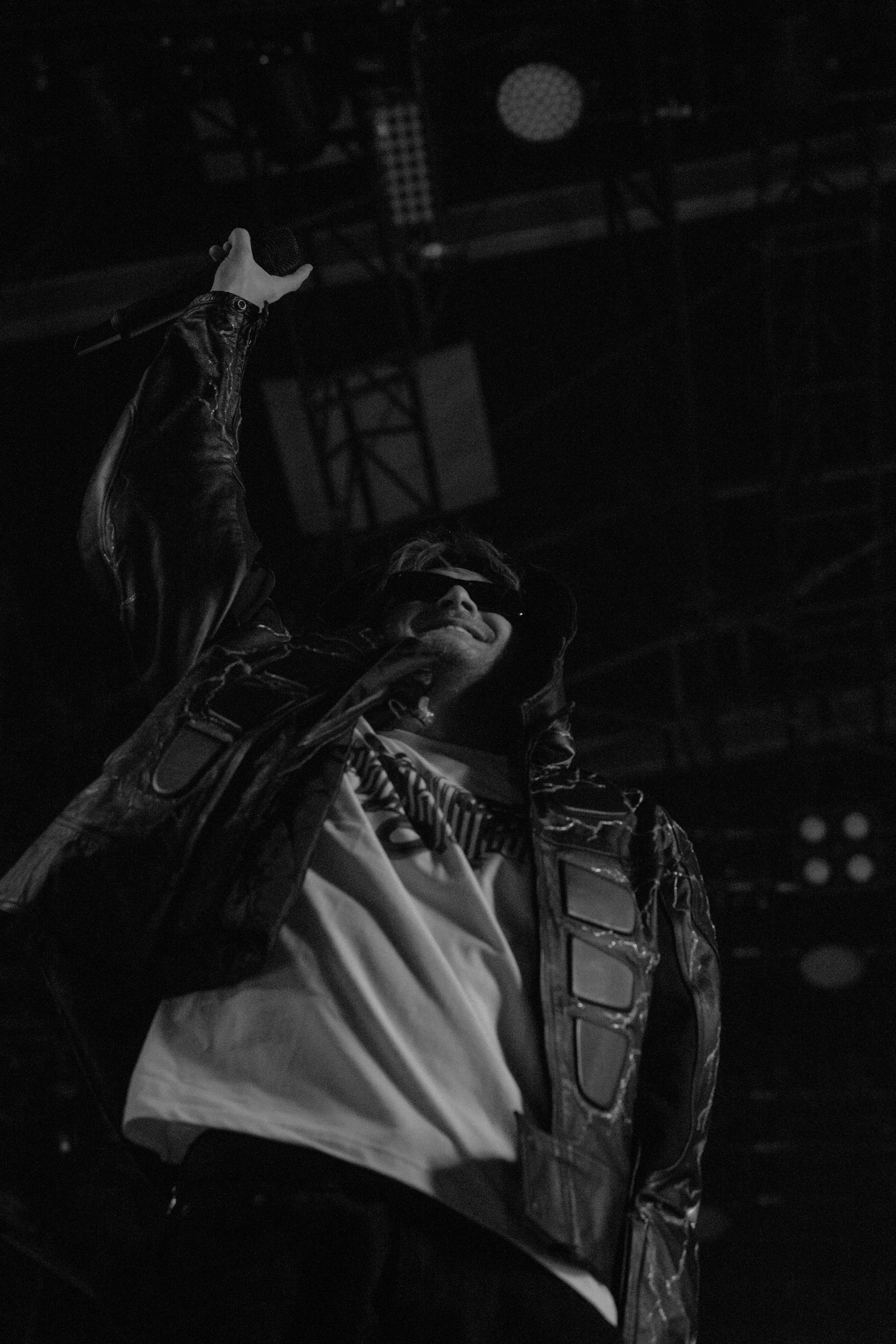 Un artiste sur scène portant des lunettes de soleil, un blouson en cuir et un t-shirt, tenant un micro dans la main, dans une ambiance sombre et énergique.