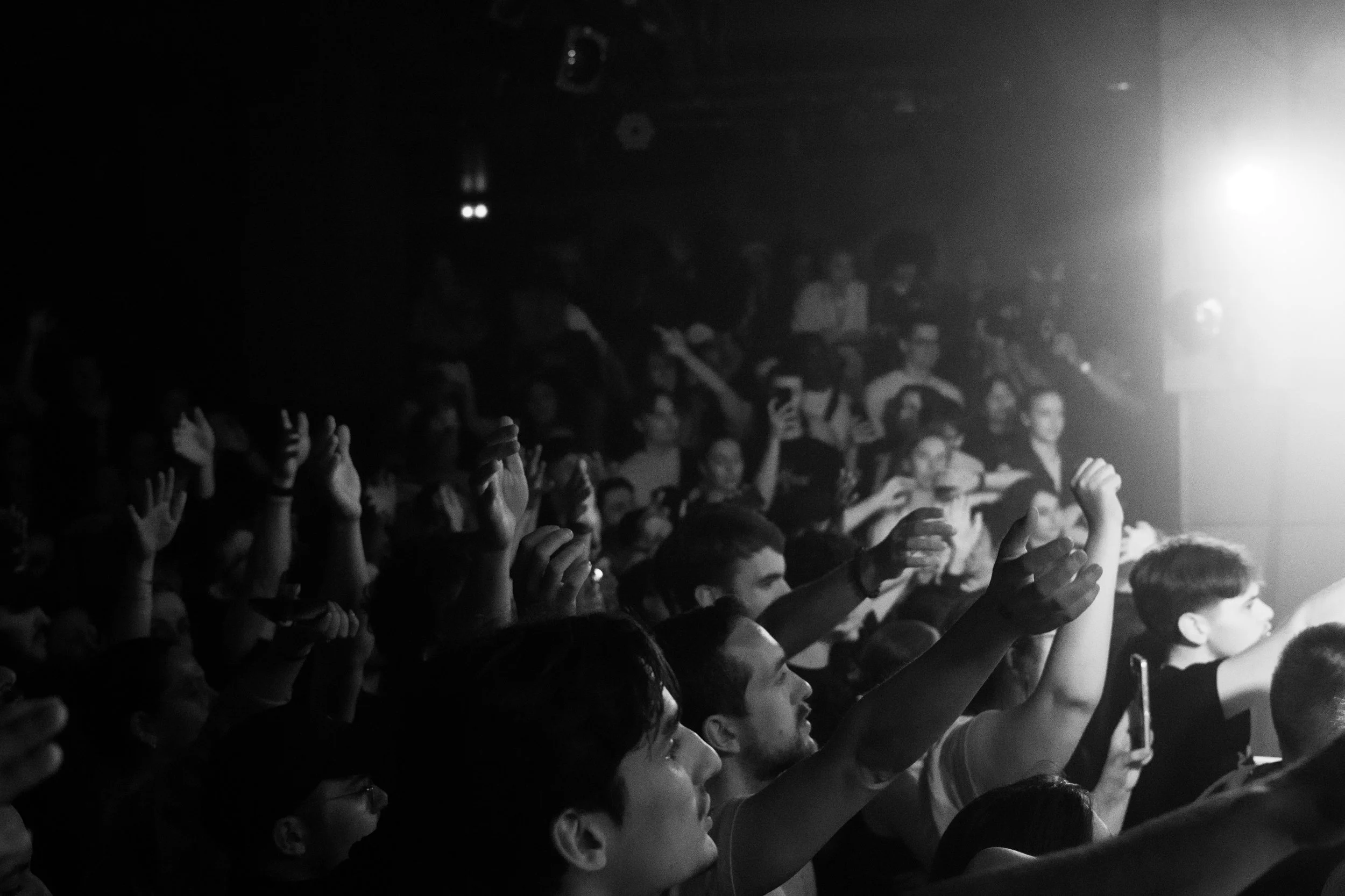Une foule de personnes dans une salle sombre, levant les bras, lors d'un concert ou d'un événement, avec une lumière forte à droite.