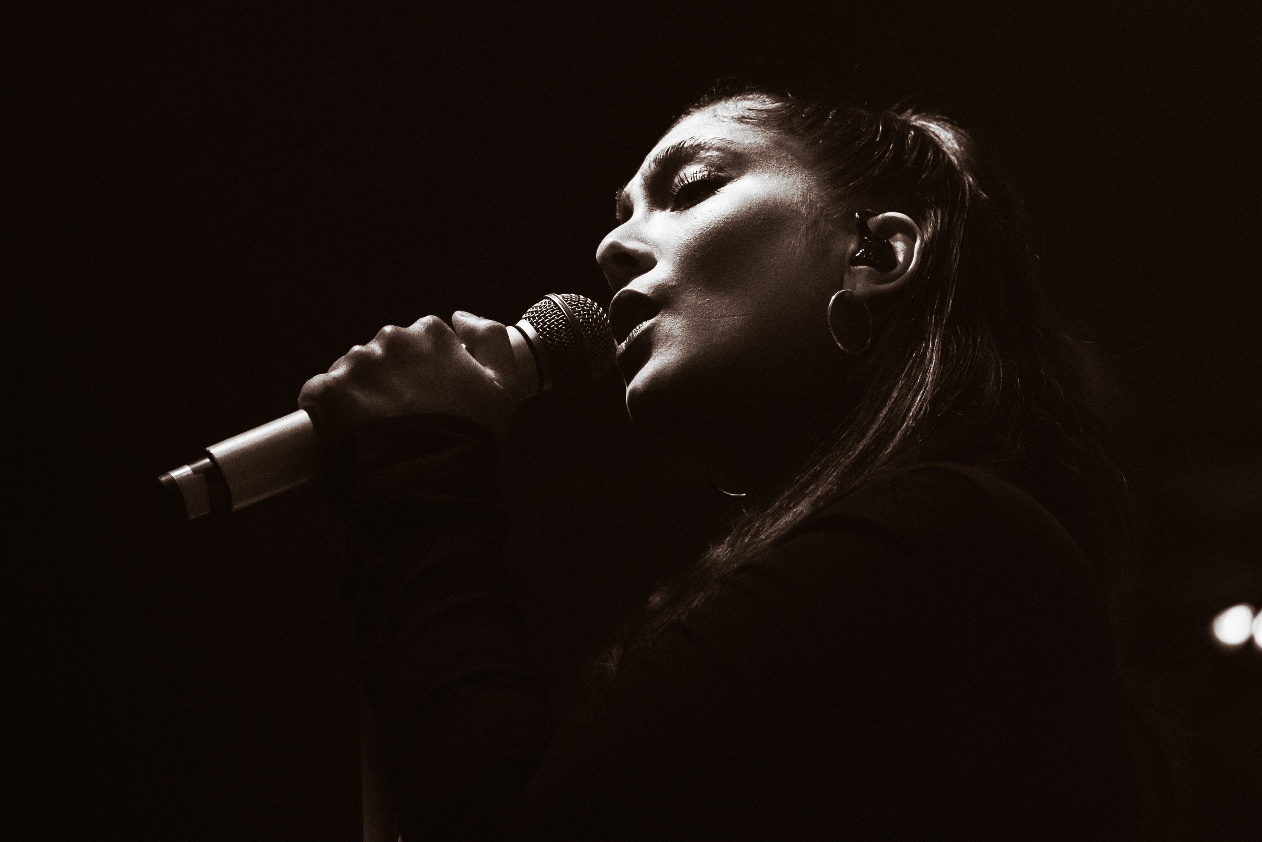 Une femme chante dans le noir, tenant un microphone près de la bouche, éclairée par une lumière douce, en noir et blanc.
