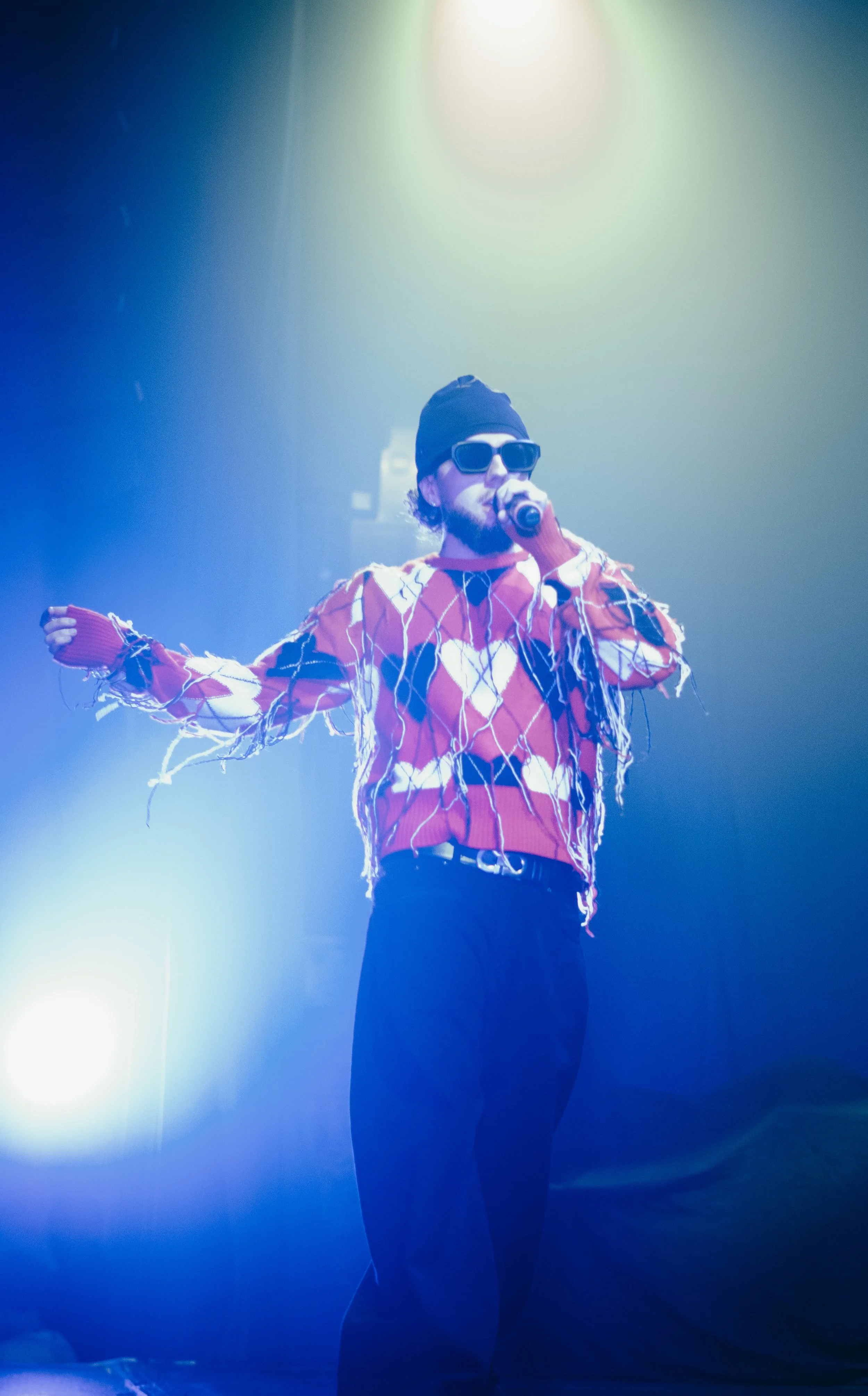 Homme portant des lunettes de soleil, un bonnet noir et un pull rouge avec des motifs, chantant ou parlant dans un microphone sur scène, entouré d'effets lumineux