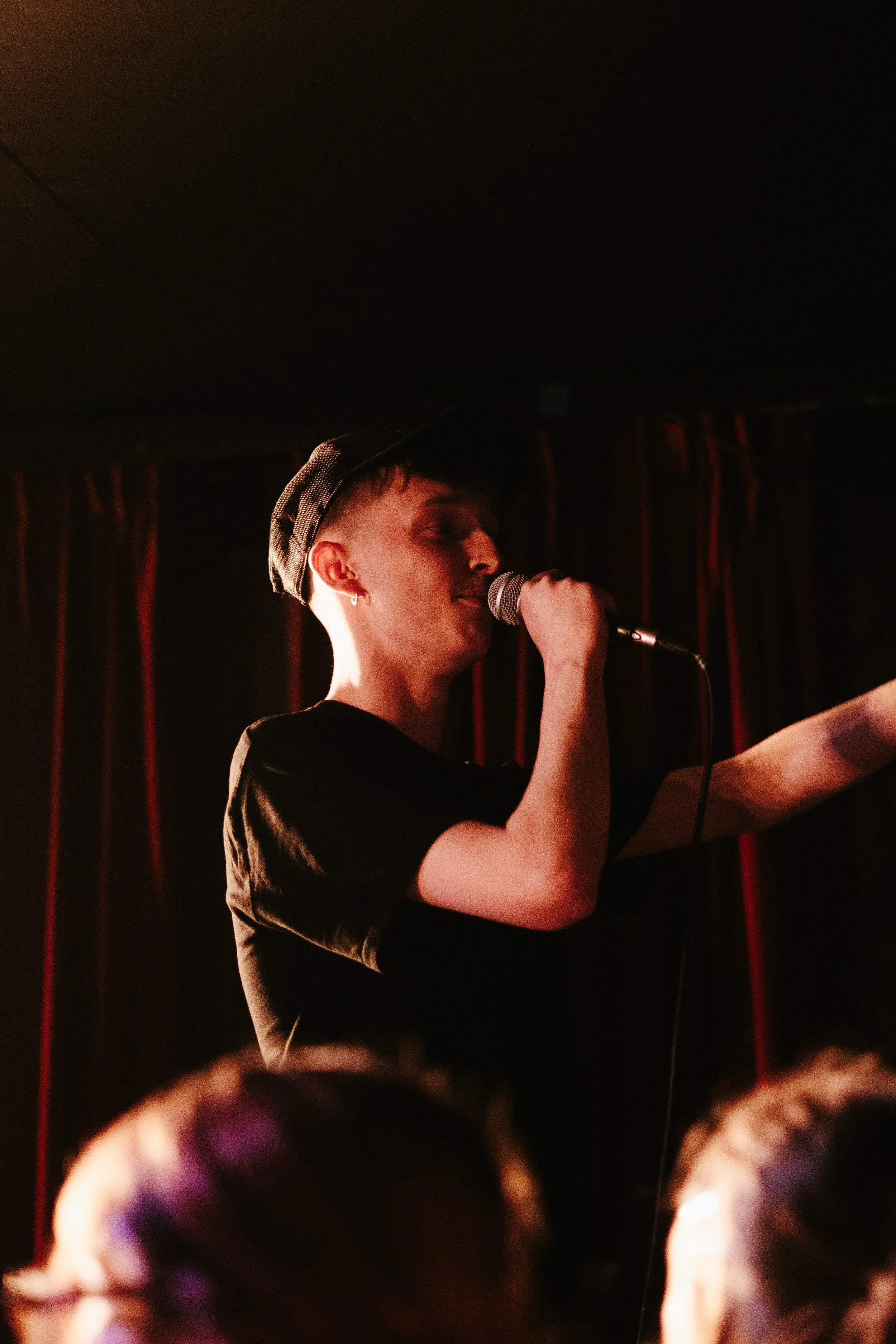 Un jeune homme portant une casquette noire chante dans un micro lors d'une performance sur scène, avec un rideau rouge en arrière-plan.