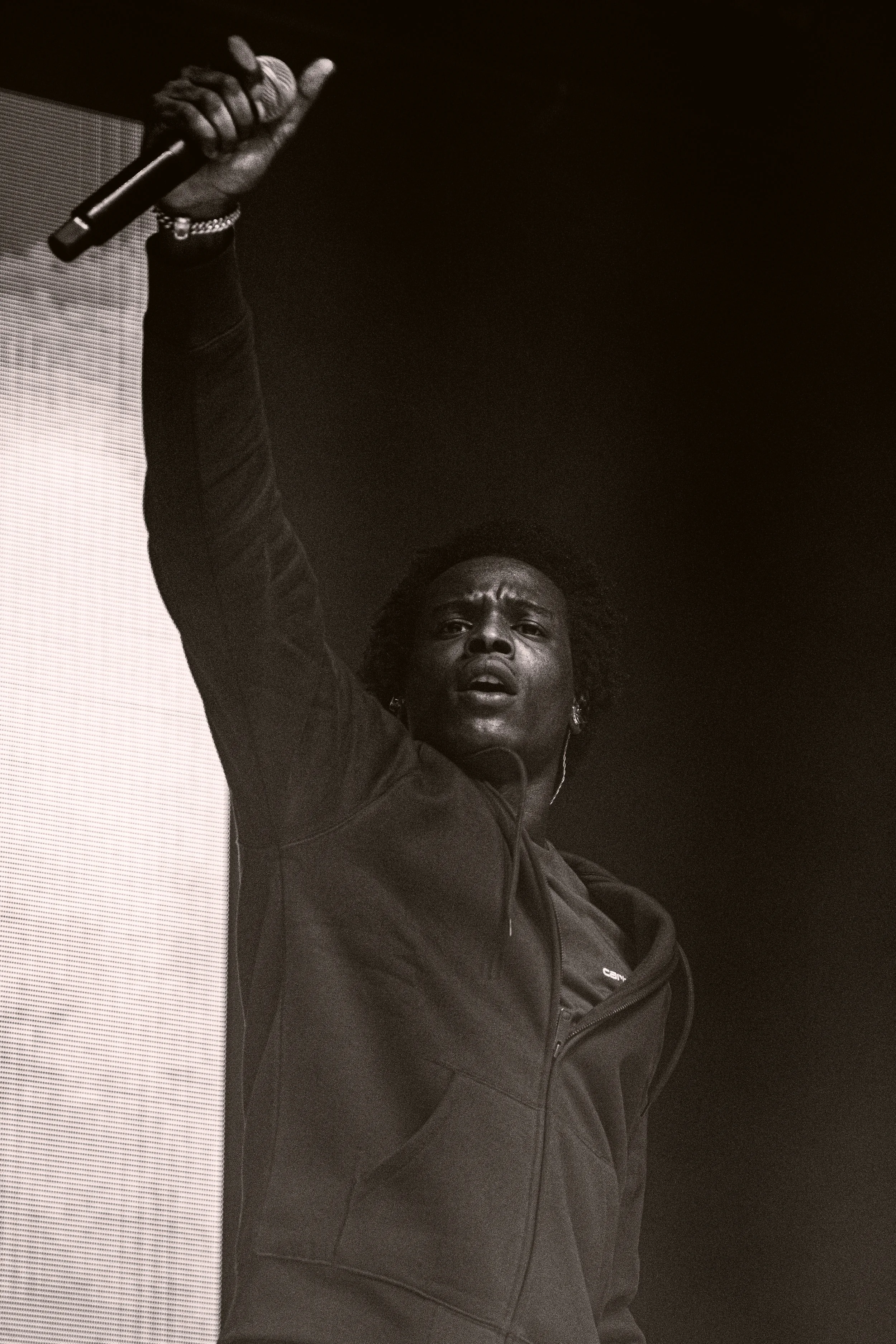 Une femme noire parle en public, levant le bras avec un micro à la main, portant une veste noire, dans un contexte of non lumineux.