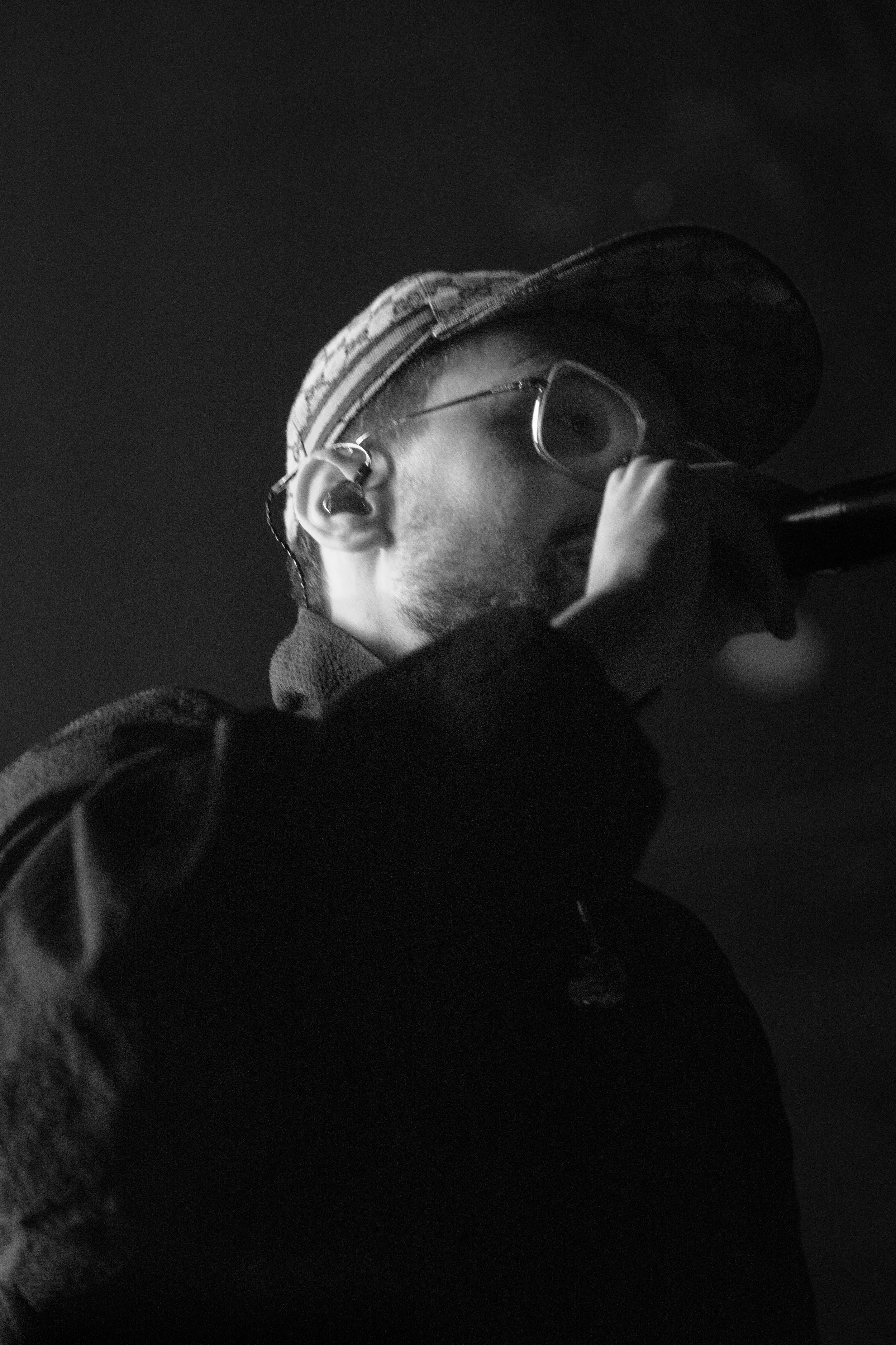 Un homme avec une casquette, des lunettes et un écouteur, en train de chanter ou de parler dans un micro, en noir et blanc.