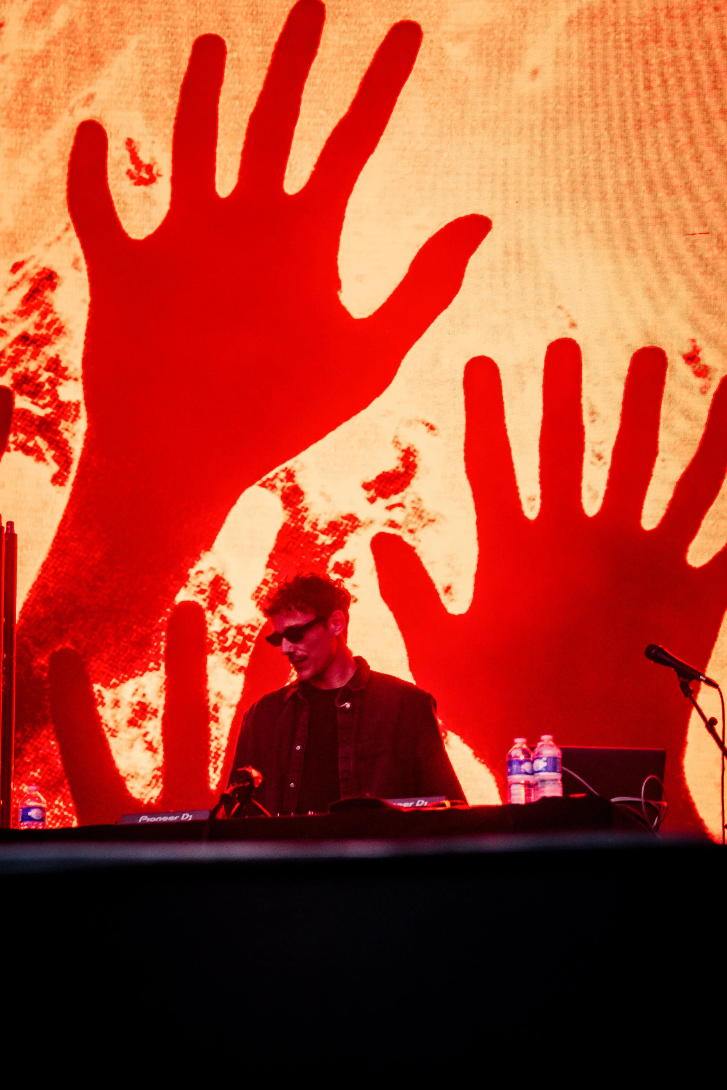 DJ en chemise noire et lunettes noires sur scène avec arrière-plan d'une projection rouge avec des mains en silhouette