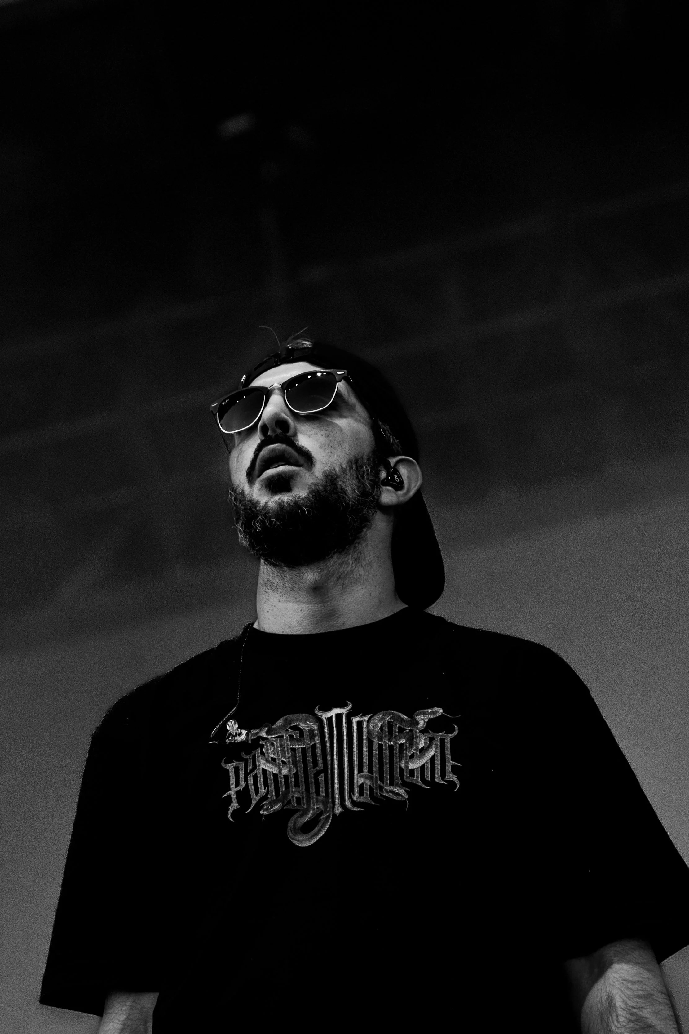 Un homme avec une barbe porte des lunettes de soleil et un t-shirt noir avec un logo complexe, semble être en extérieur dans un style noir et blanc.