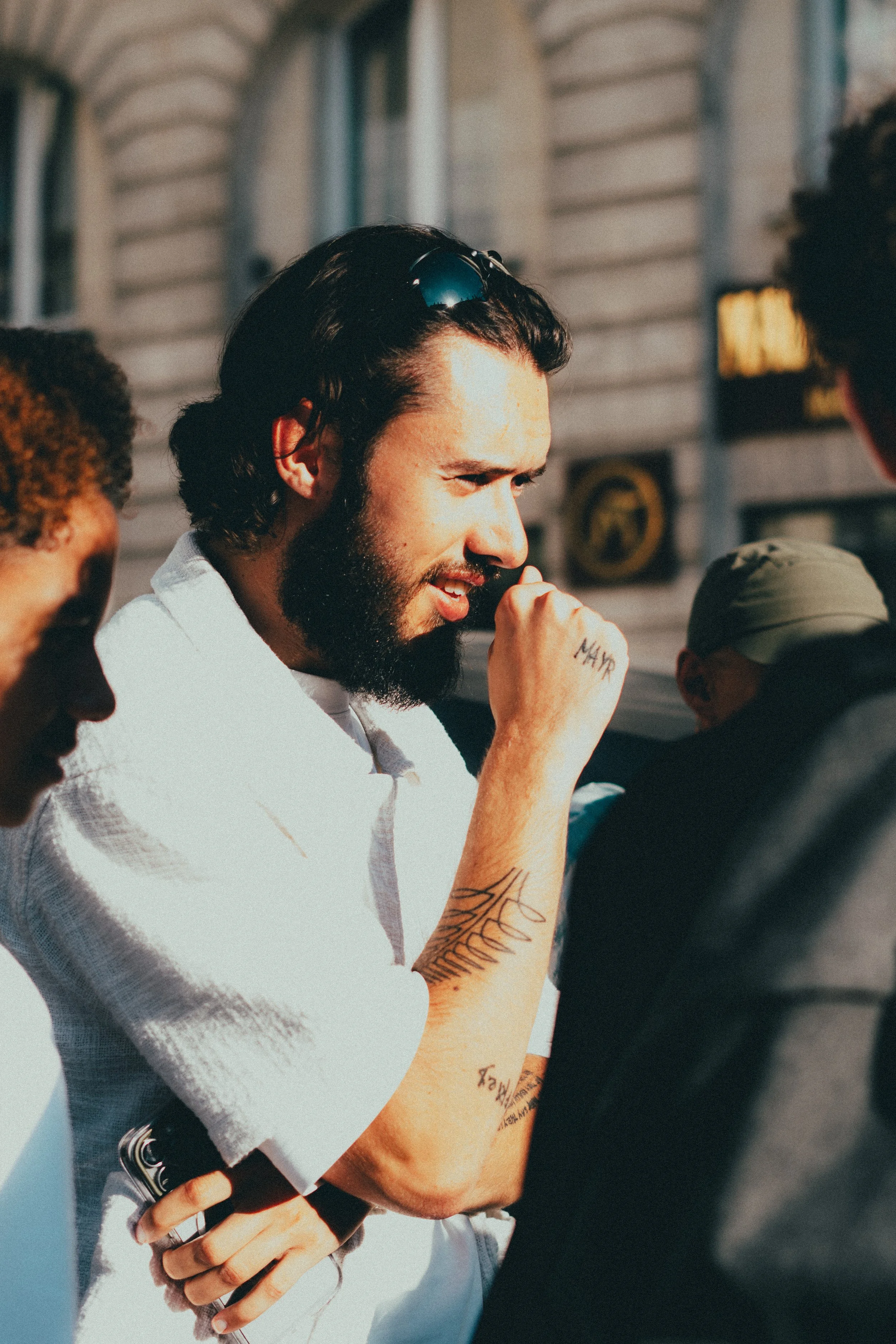 Homme avec barbe et tatouages, portant des lunettes de soleil sur la tête, parlant à un groupe, en extérieur, bâtiments en arrière-plan.