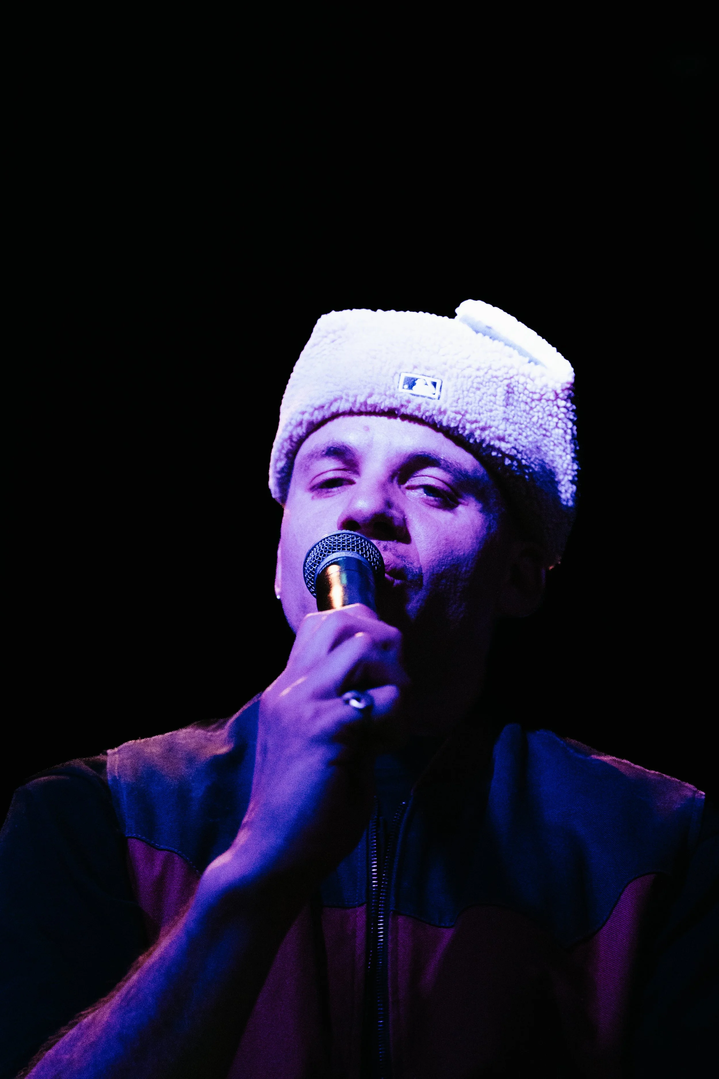 Un homme chante dans un microphone, portant un bonnet blanc et une veste noire, sous un éclairage violet sur un fond noir.