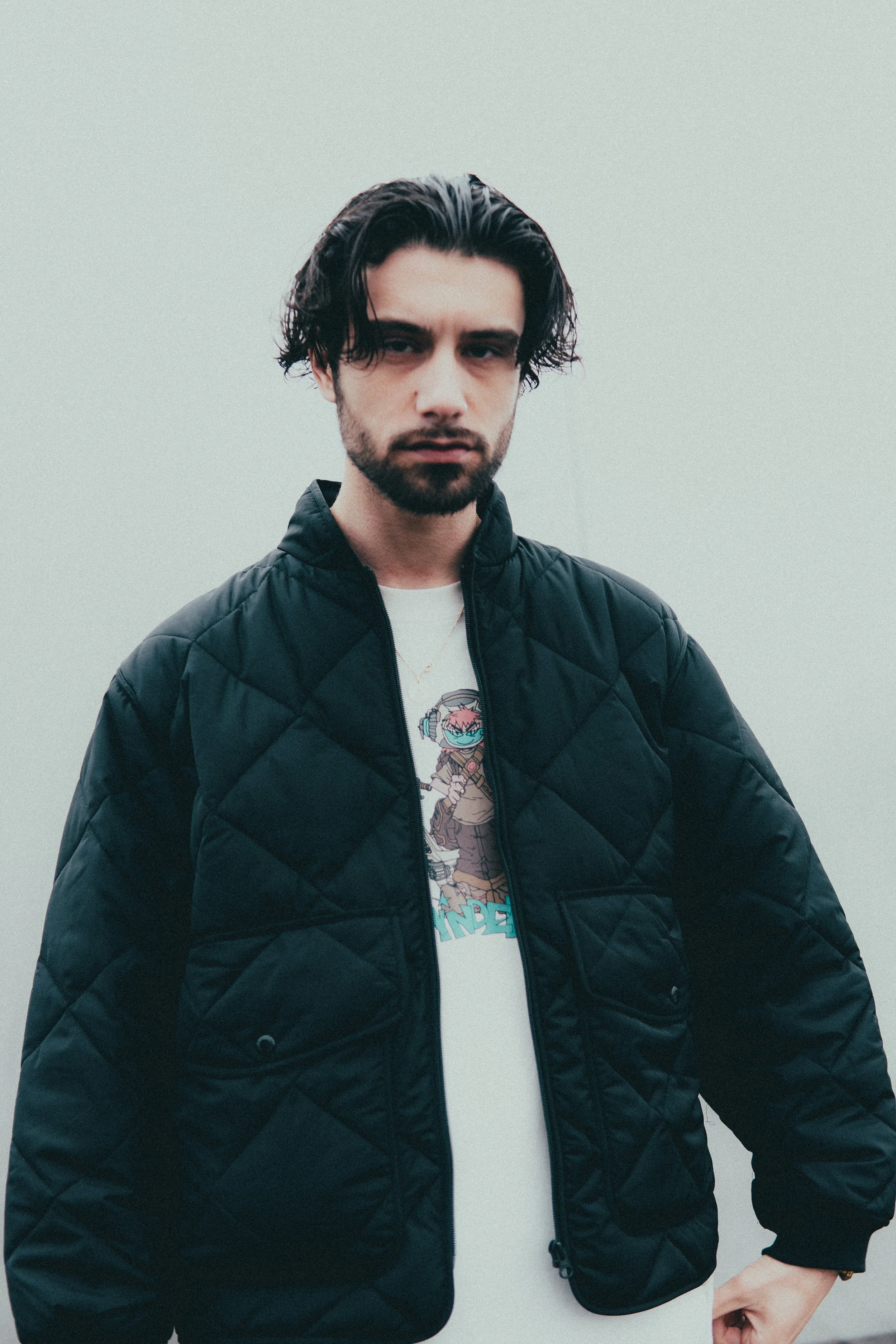 Jeune homme avec cheveux longs et barbe portant une veste noire en quilt et un t-shirt avec un personnage de dessin animé