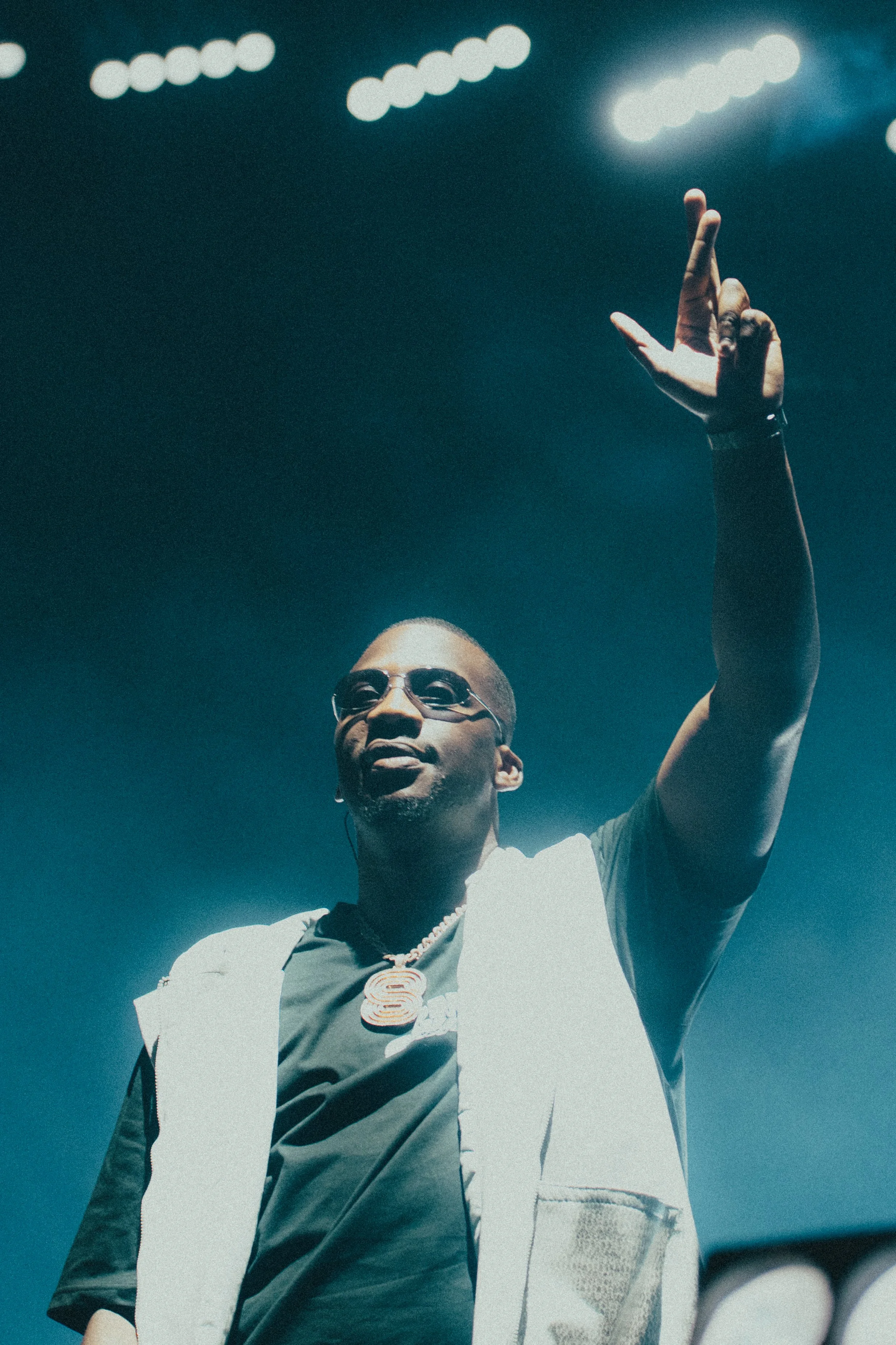 Une personne portant des lunettes de soleil et un collier, levant la main lors d'un concert ou événement en intérieur avec un éclairage de scène.