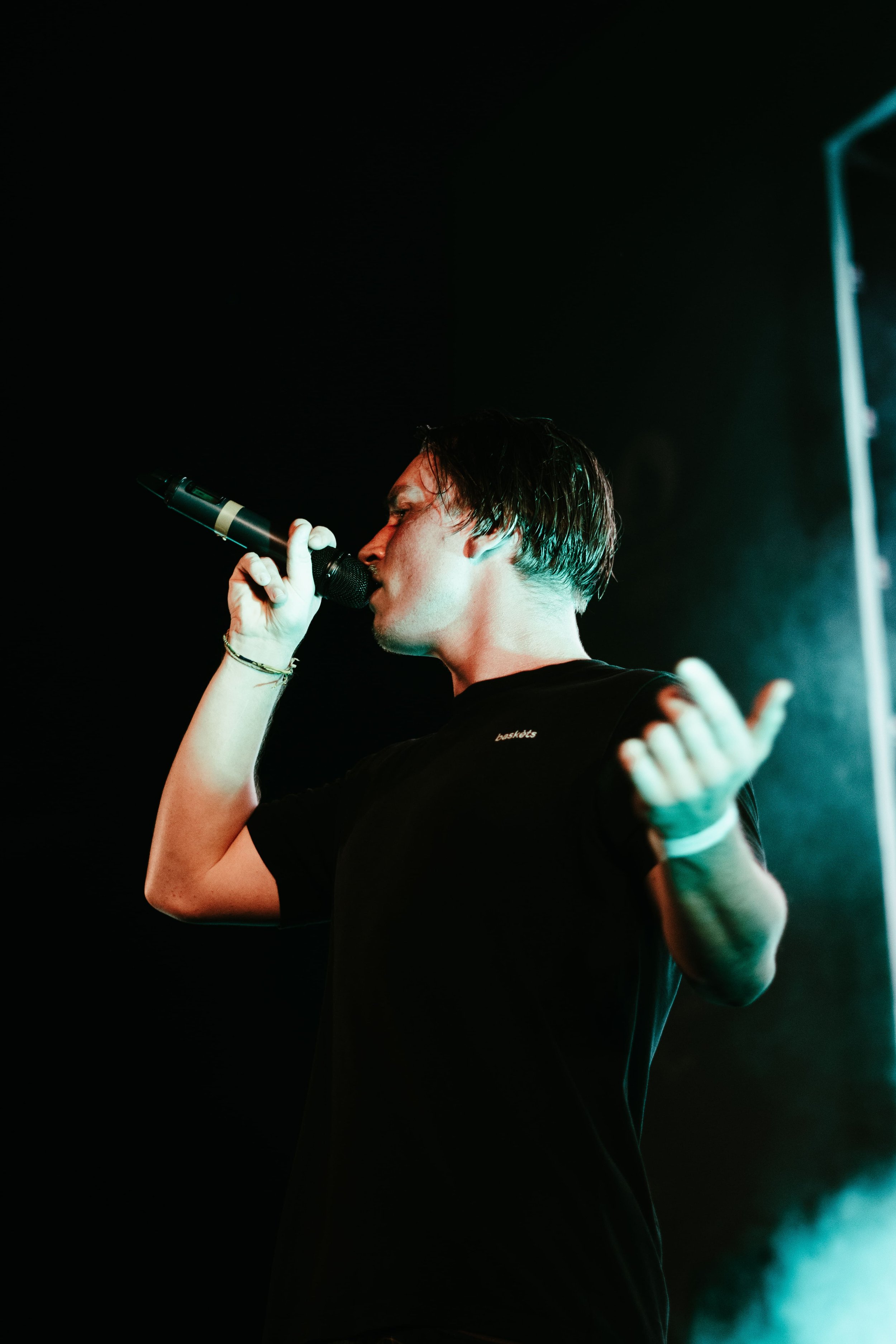 Un jeune homme chante dans un microphone lors d'une performance, vêtu d'un t-shirt noir avec un éclairage de scène coloré.