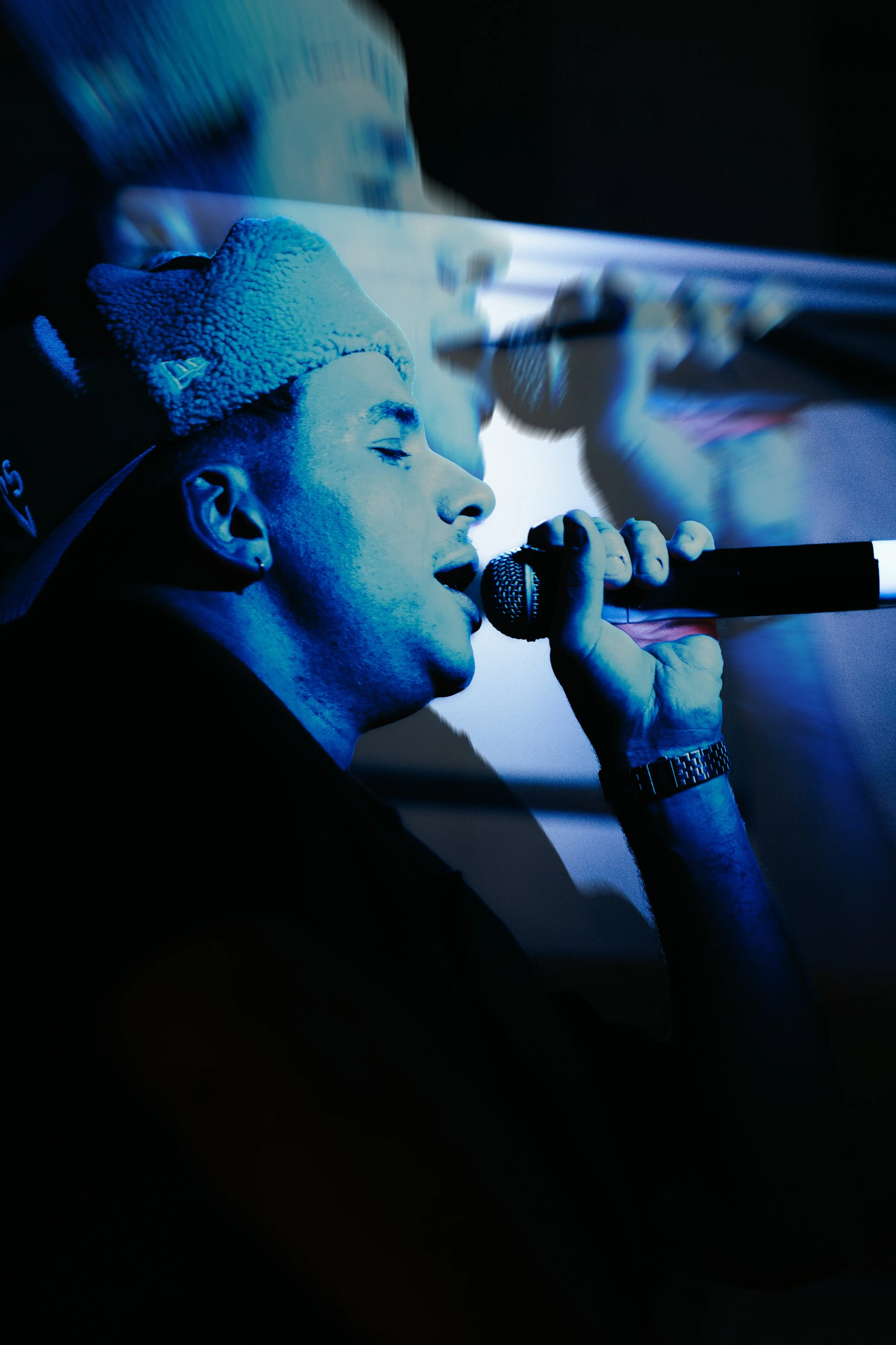 Jeune homme chantant dans un micro sous un éclairage bleu, portant un bonnet en peluche et une montre.