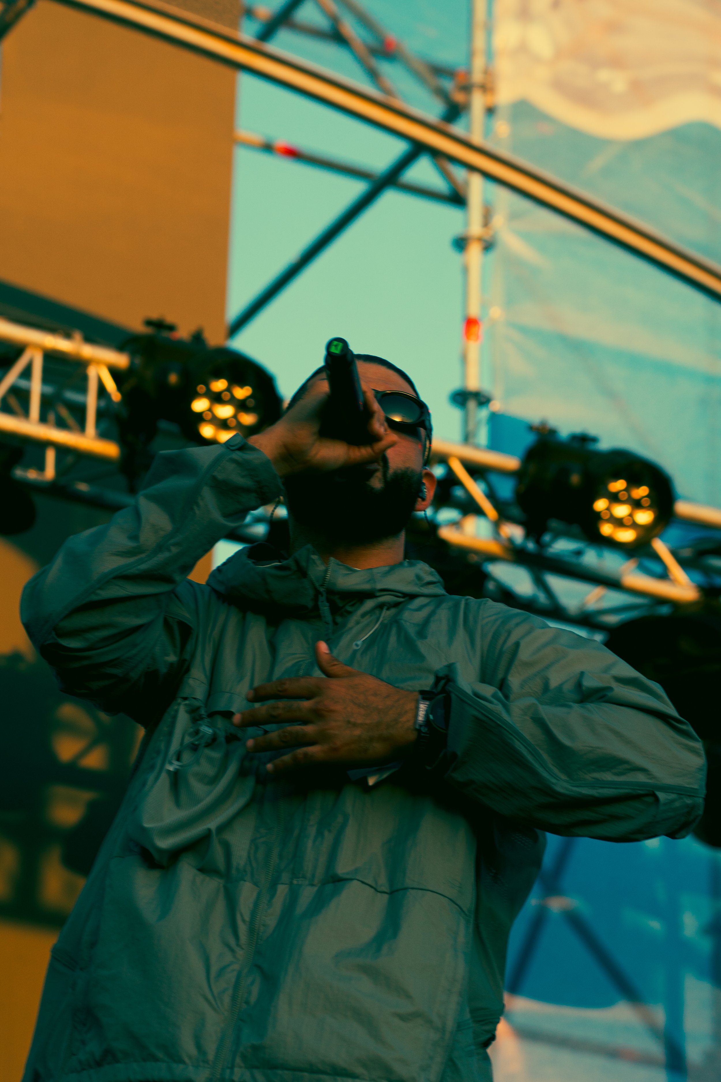 Un homme portant une veste grise, des lunettes de soleil, chantant ou parlant dans un micro lors d'un événement en plein air, avec des lumières de scène et une structure métallique en arrière-plan.