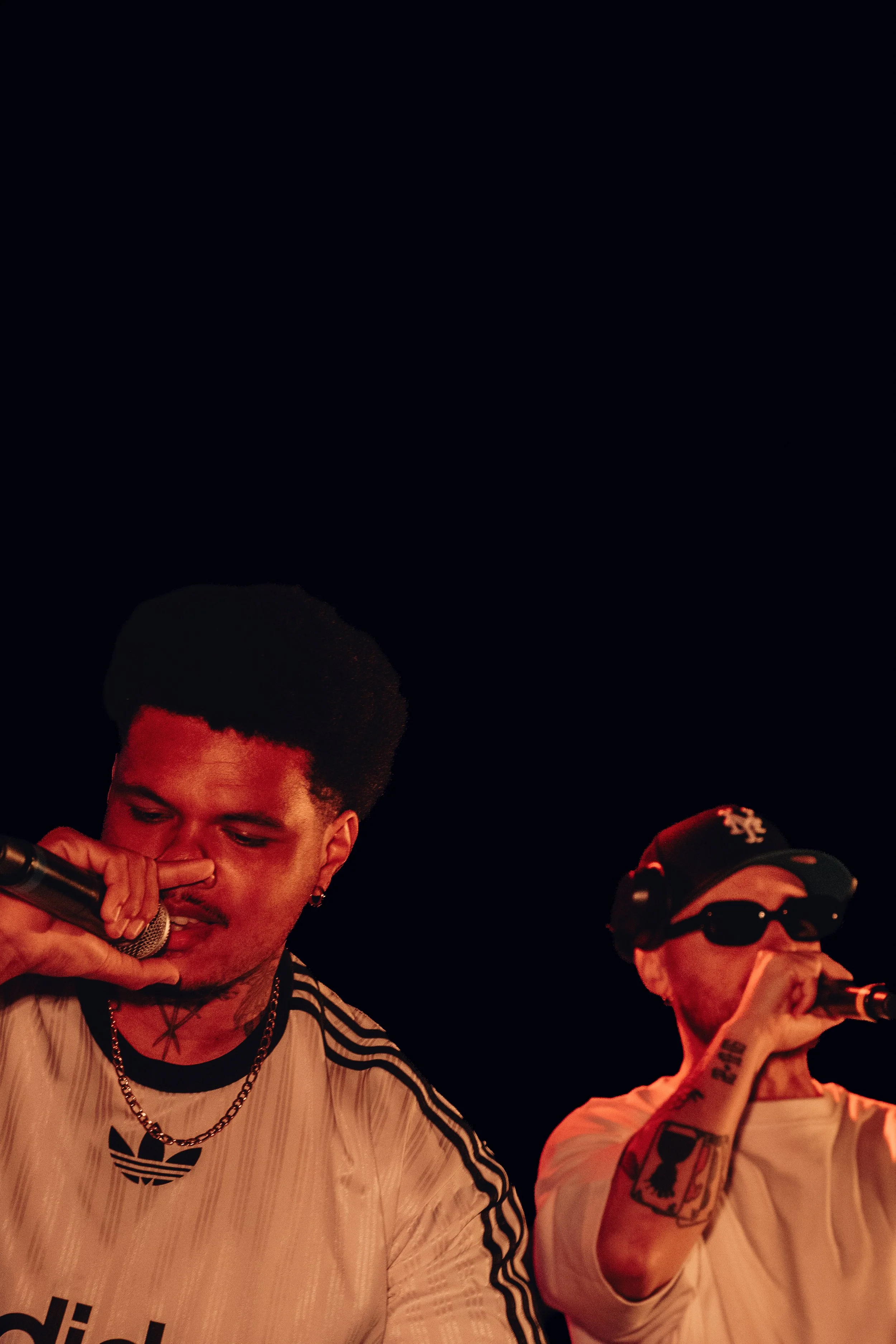 Deux jeunes hommes chantent ou parlent dans un microphone lors d'une performance. L'un porte un t-shirt blanc avec un logo Adidas, des chaînes et a une coupe de cheveux afro. L'autre porte des lunettes de soleil, un bonnet et un t-shirt blanc. L'ambi