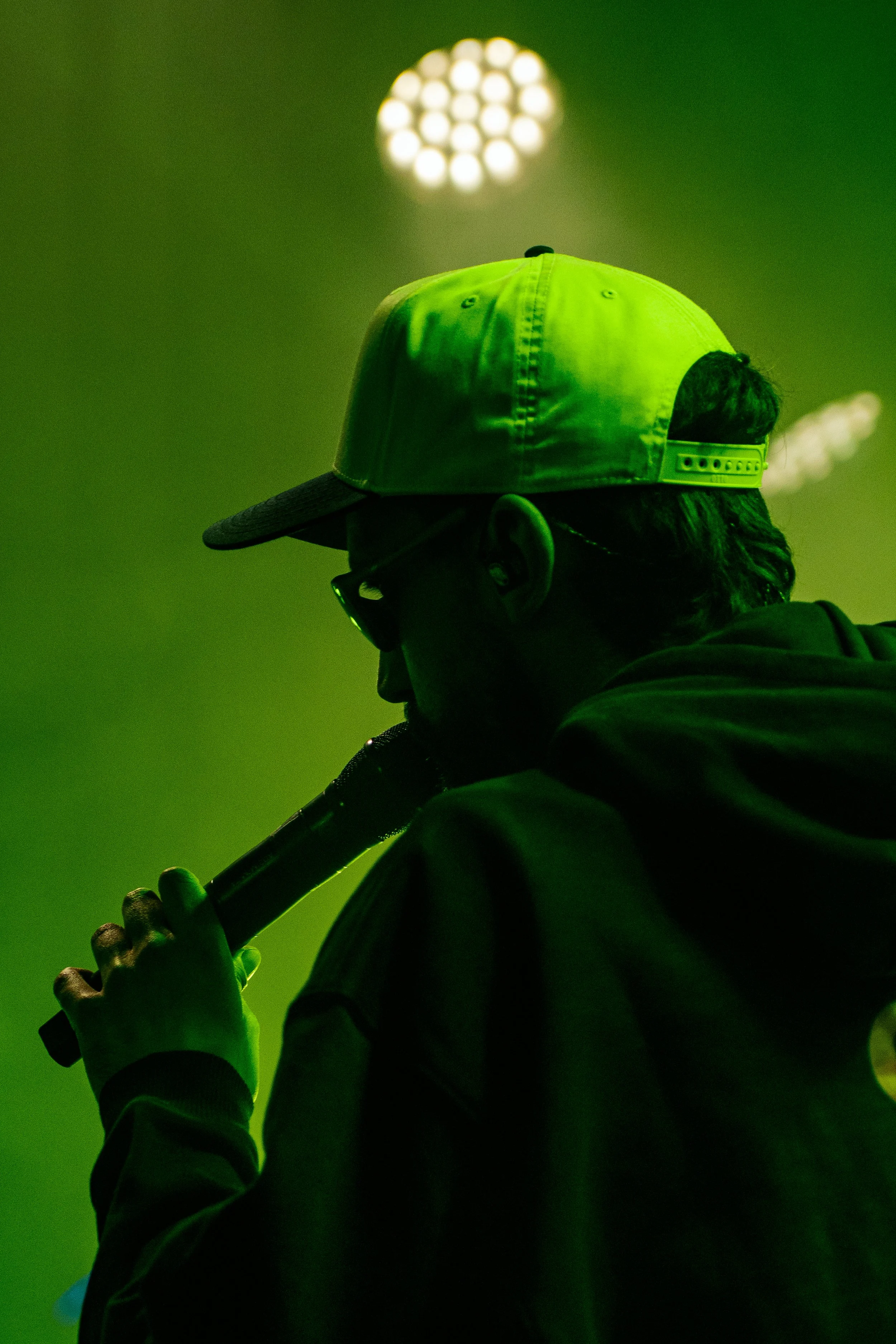 Un homme portant une casquette verte, des lunettes de soleil, et une veste, tenant un microphone près de la bouche dans une ambiance sombre et verte.