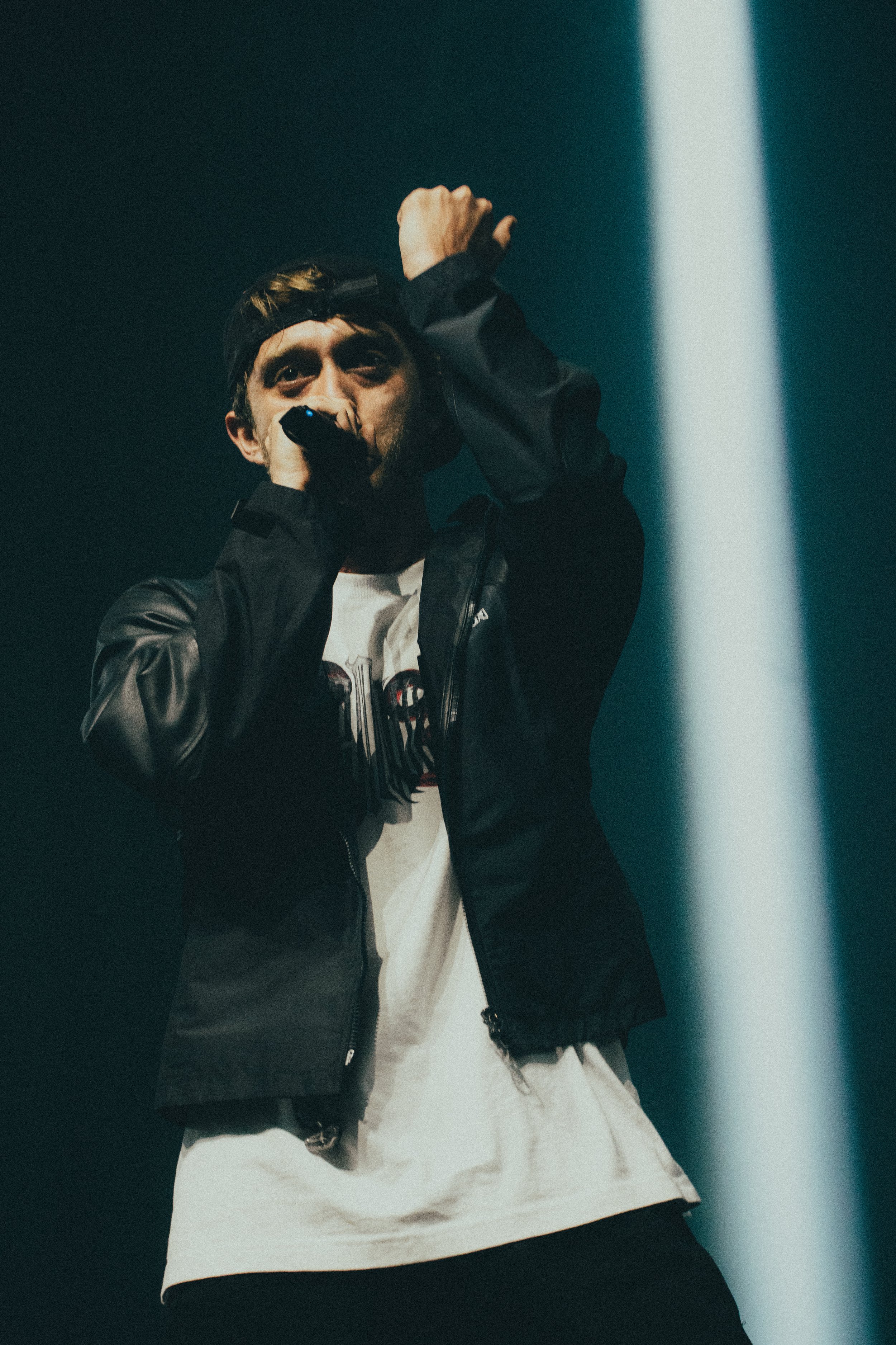 Un homme en tenue décontractée, portant une veste noire et un t-shirt blanc, chante avec un micro, éclairé par un faisceau lumineux sur scène.