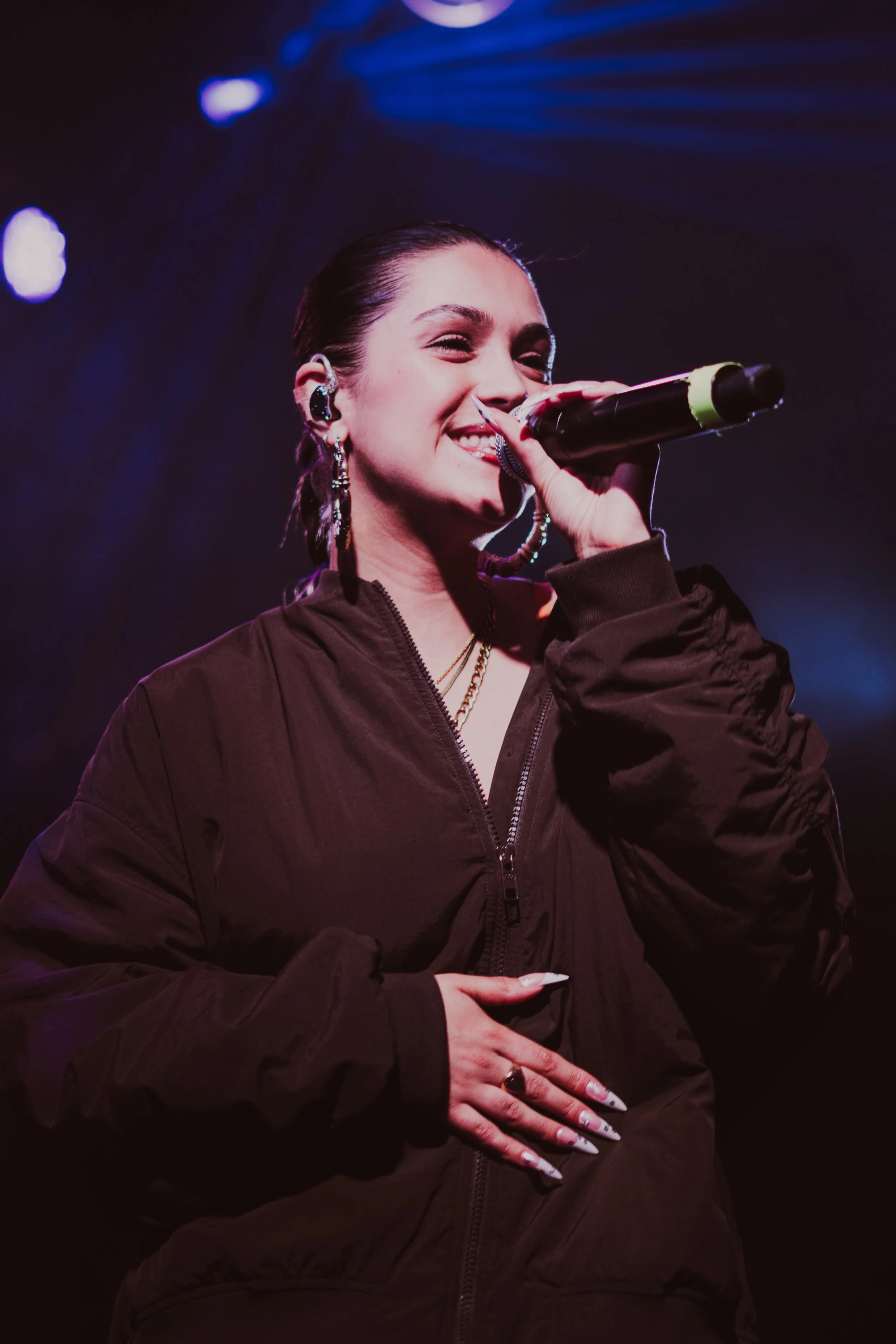 Une femme chante sur scène avec un microphone, portant une veste noire et des bijoux. Elle sourit en chantant.