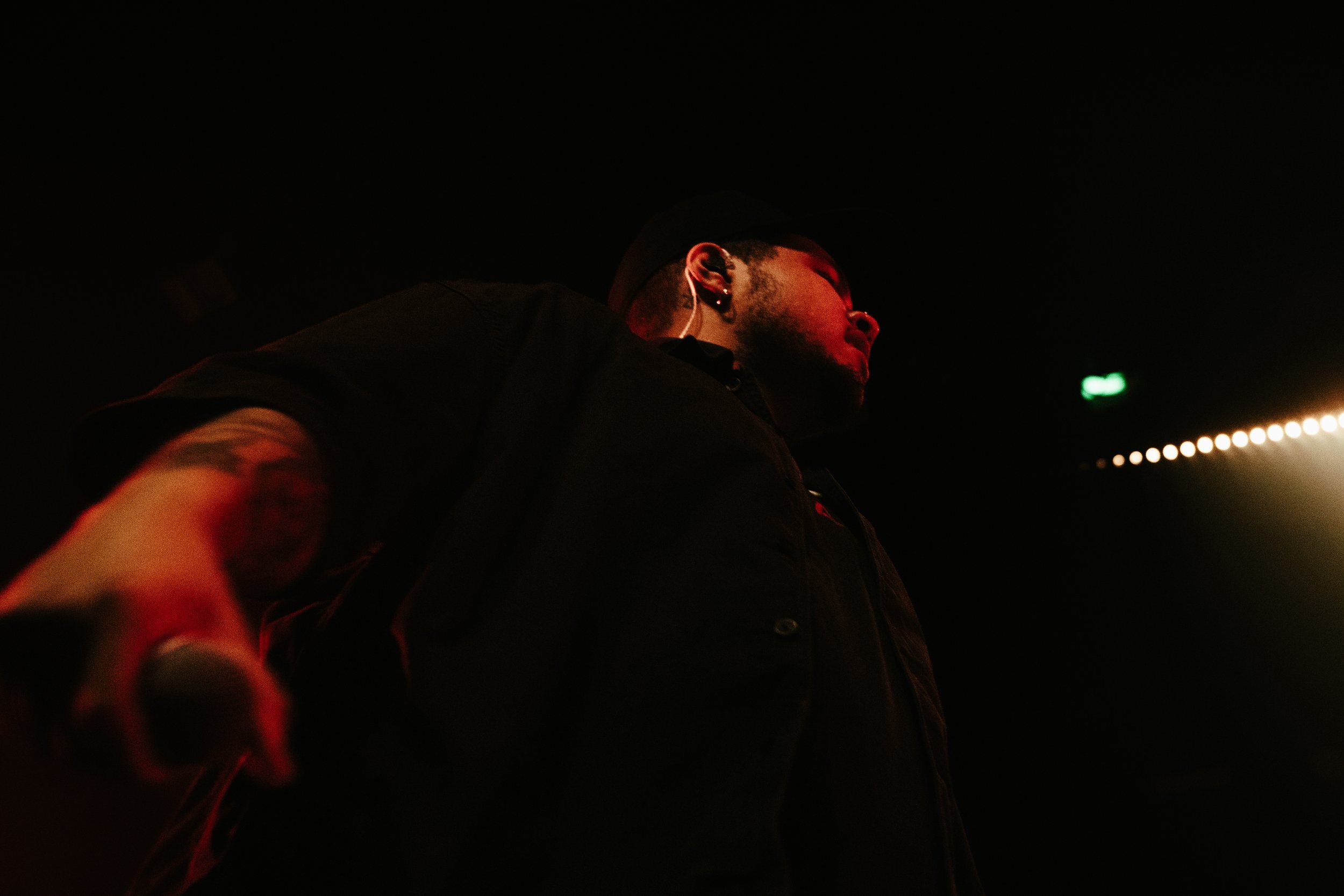 Homme dans un environnement sombre, portant une veste noire, portant des écouteurs, vu de côté, éclaire par un faisceau lumineux pointe vers la droite.