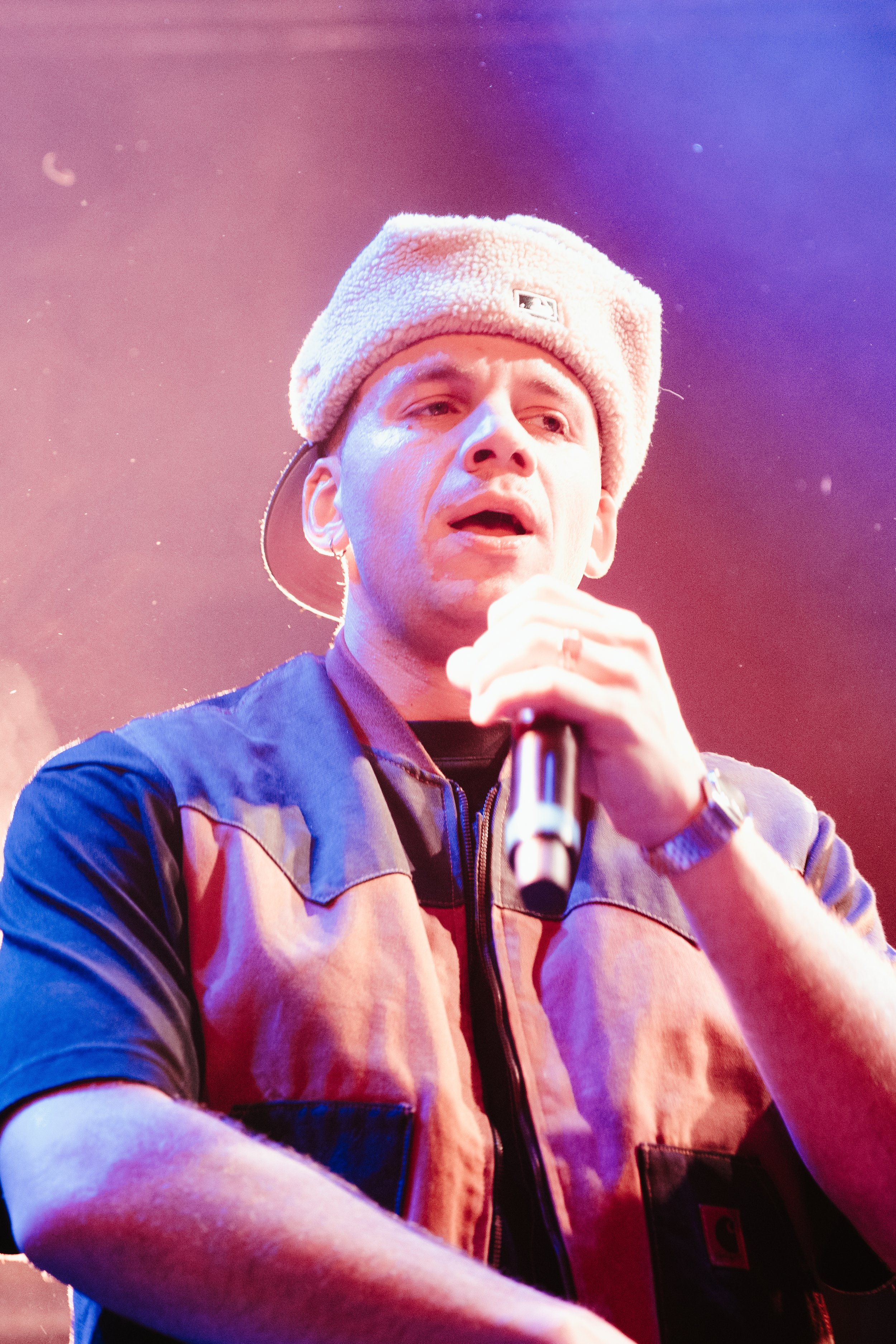 Un homme chante ou parle en tenant un microphone, portant un bonnet en tricot et une veste de couleur beige et noire.