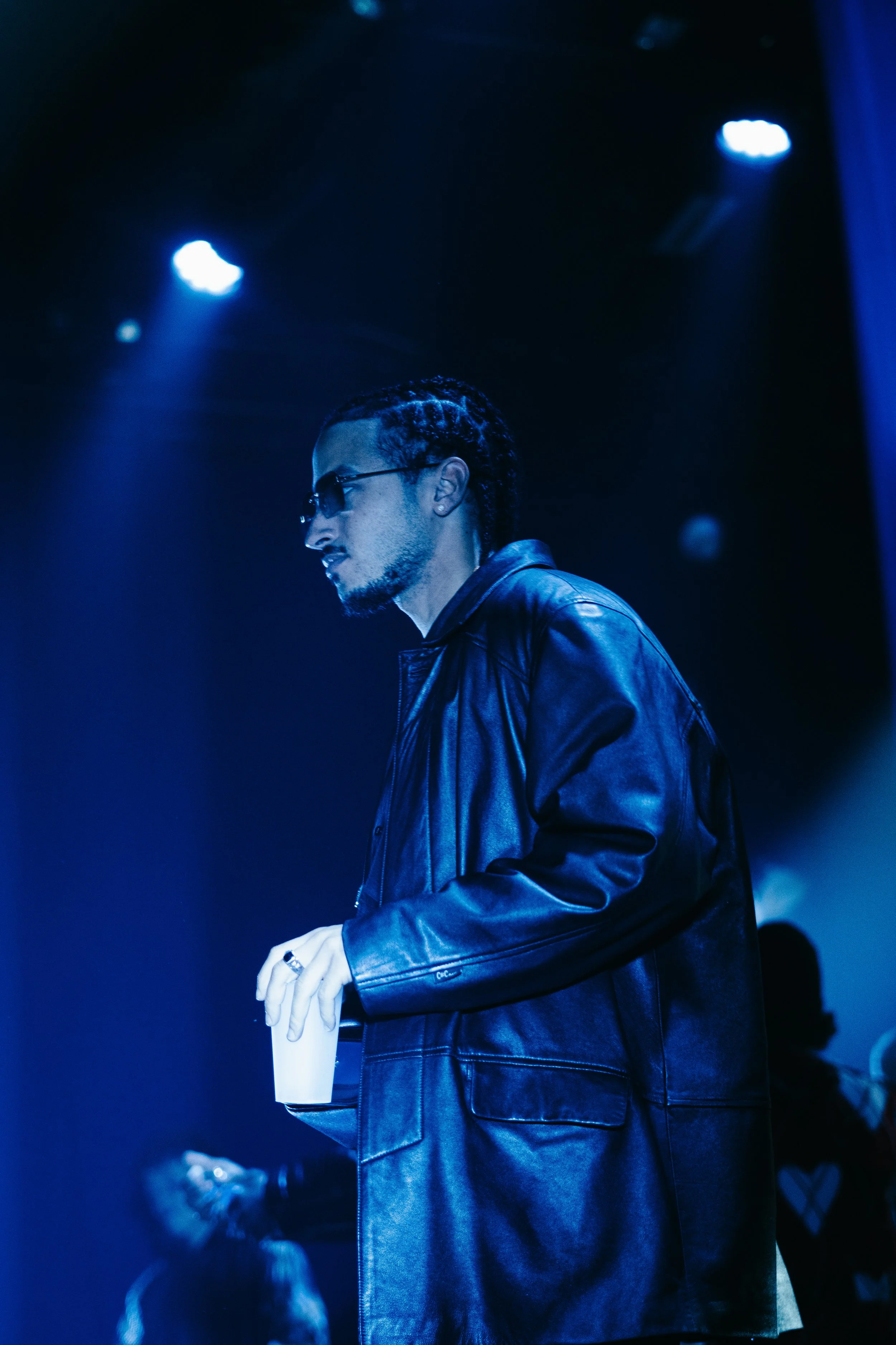 Un homme portant un manteau en cuir noir et des lunettes de soleil tient une tasse blanche dans une ambiance sombre avec un éclairage bleu.
