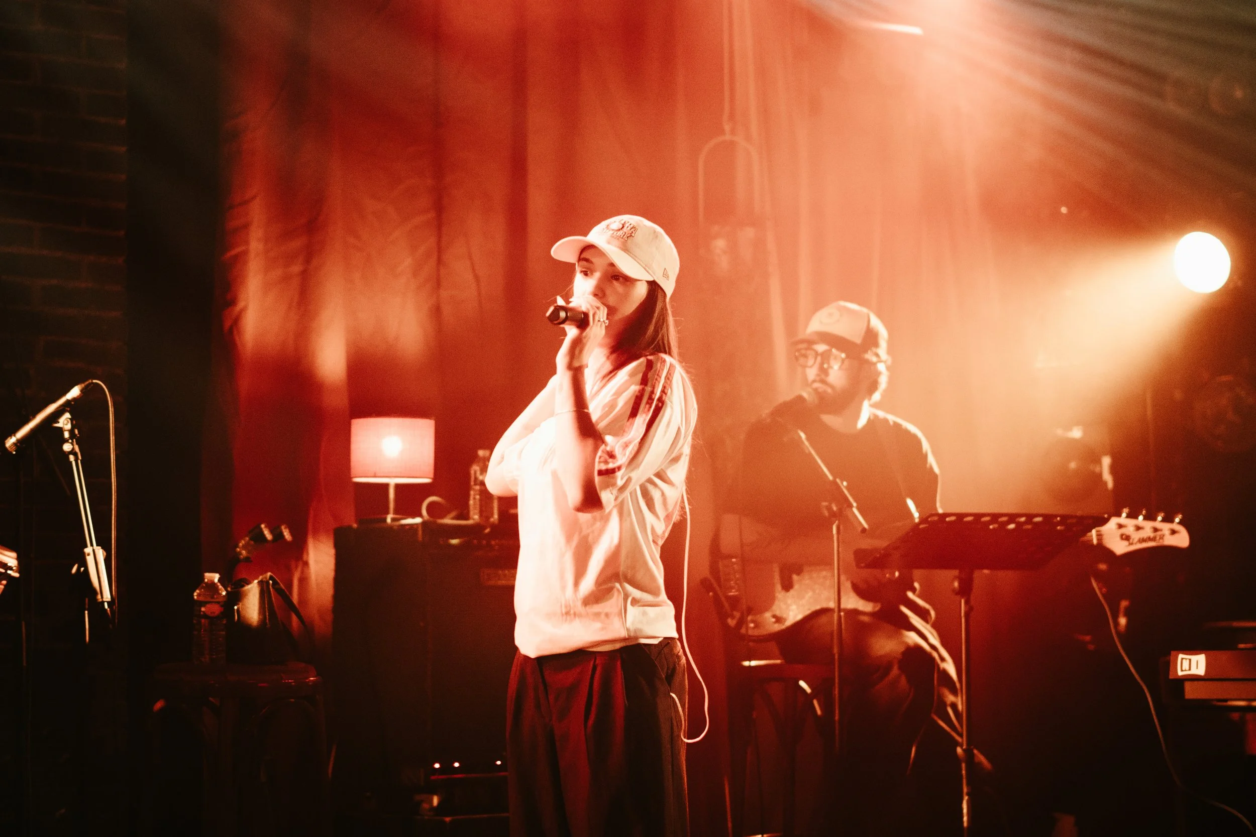 Une femme chante avec un micro dans un environnement sombre et chaud, accompagnée d'un musicien jouant de la guitare électrique, sous un éclairage doux et ambiant.