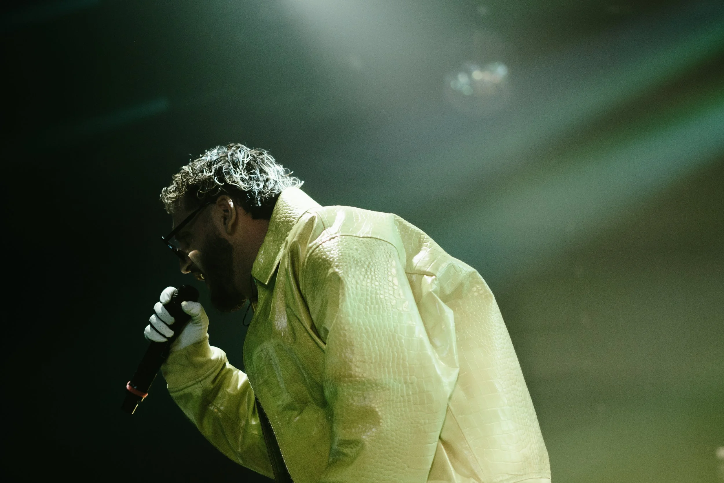 Un homme chantant en microphone, portant une veste jaune phosphorescente, lunettes et gants, sous un éclairage vert