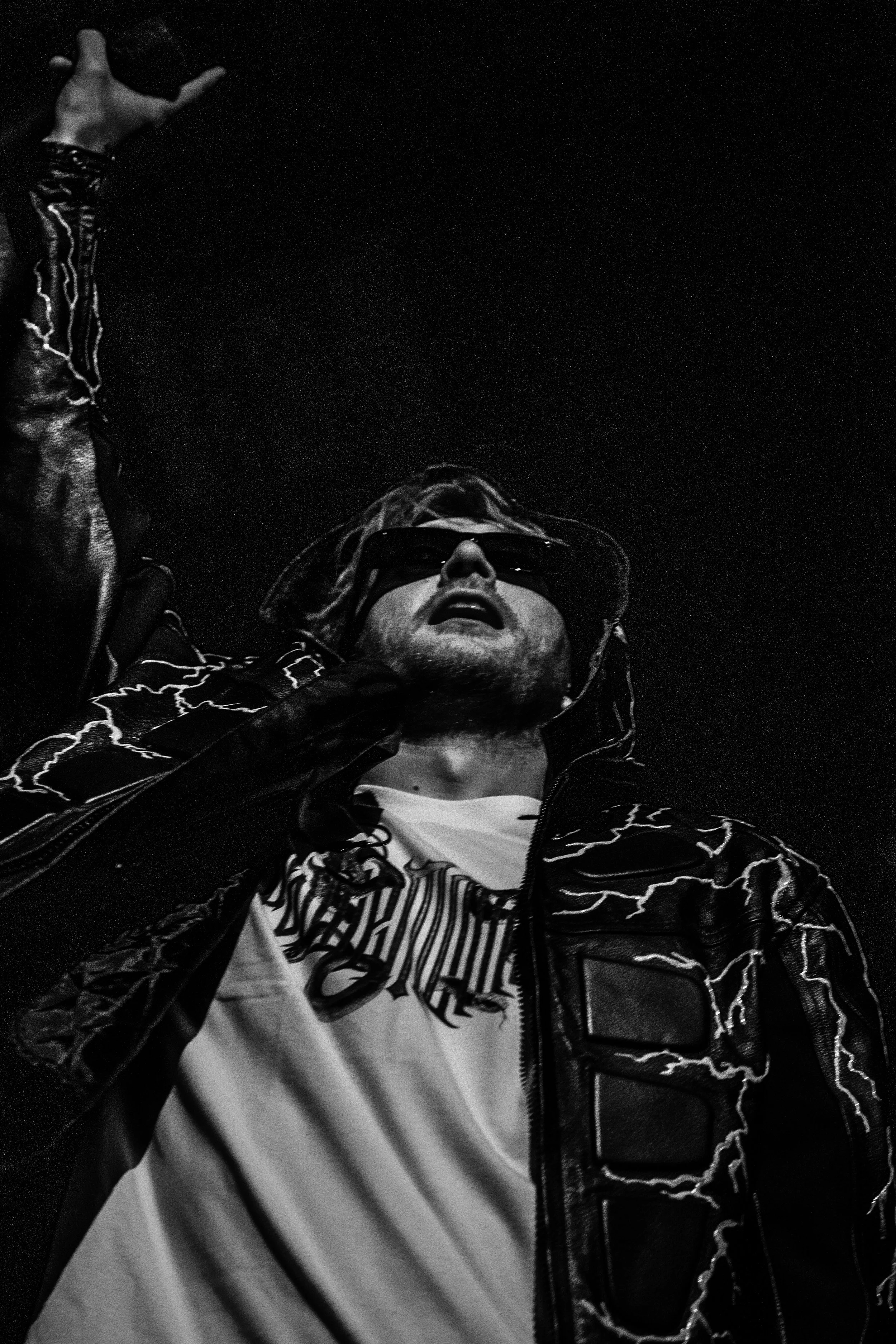 Un homme portant des lunettes de soleil, une veste noire avec des motifs de foudre, et un t-shirt blanc avec une inscription, regardant vers le haut dans une photographie en noir et blanc.