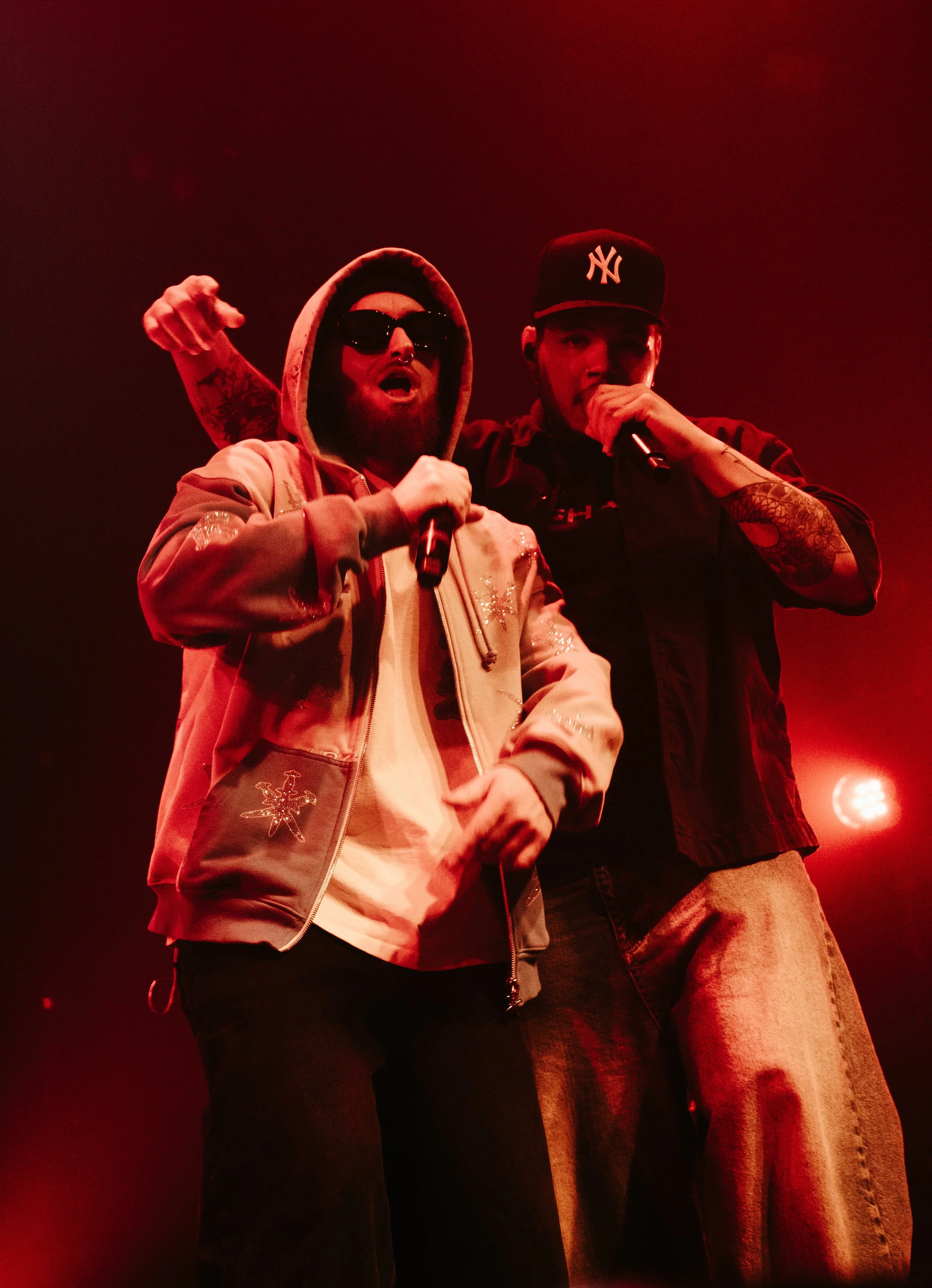 Deux rappeurs sur scène sous un éclairage rouge, tenant des microphones, avec un verre à la main, l'un portant une casquette New York et des vêtements décontractés, l'autre portant des lunettes de soleil et un hoodie.