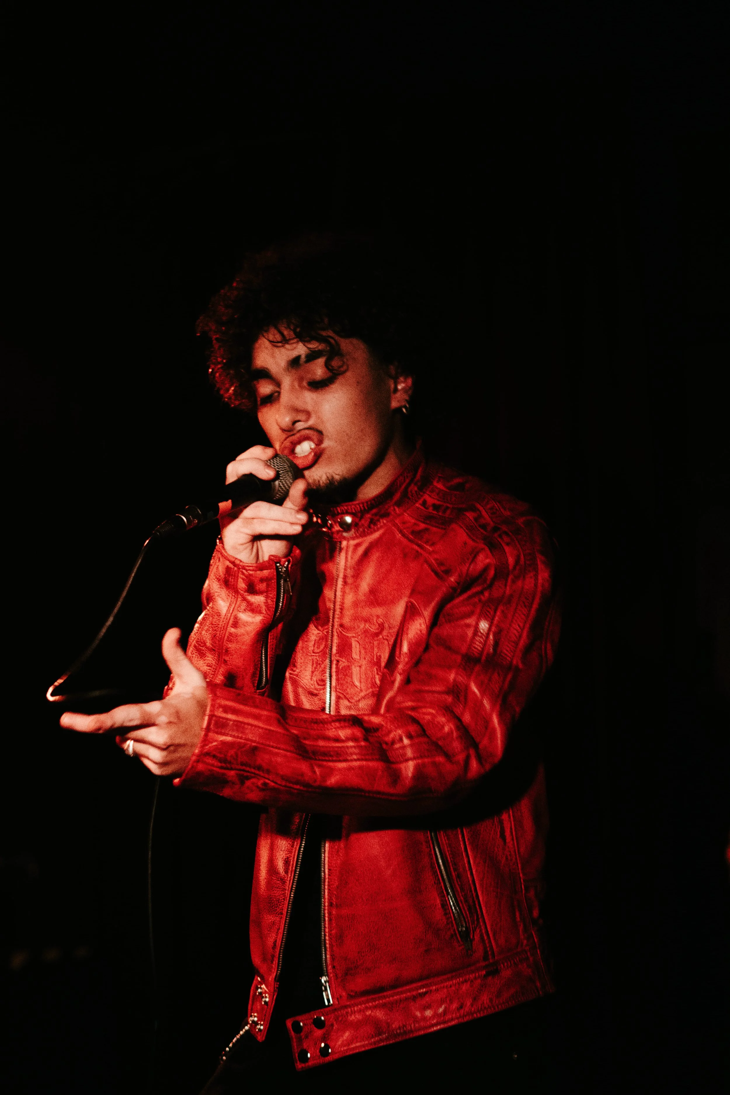 Un homme chante avec une gestuelle expressive dans un éclairage sombre, portant une veste en cuir rouge.