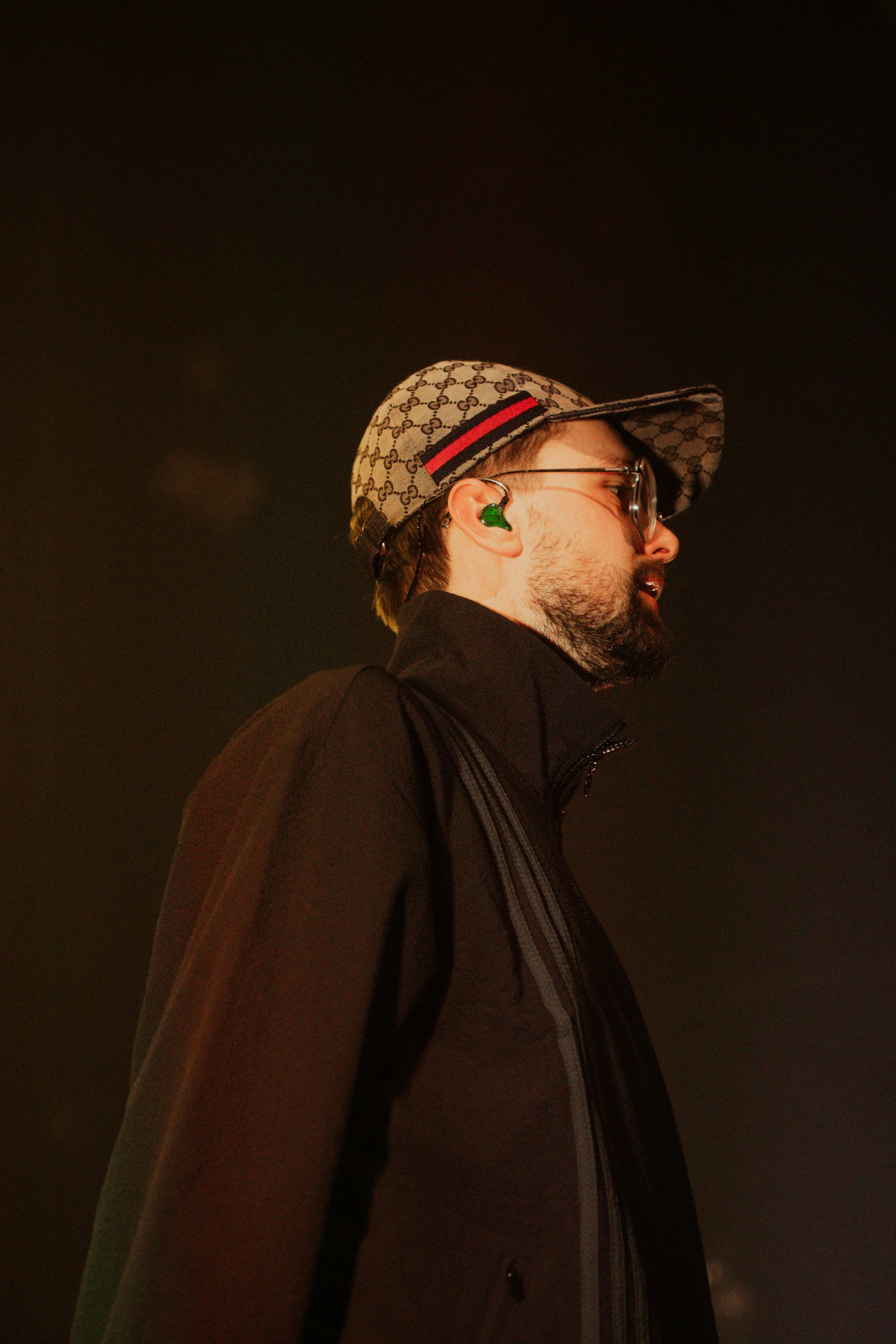 Homme portant un chapeau de baseball Gucci, lunettes, vêtements noirs, avec un écouteur vert dans l'oreille, contre un fond sombre, vue de profil.