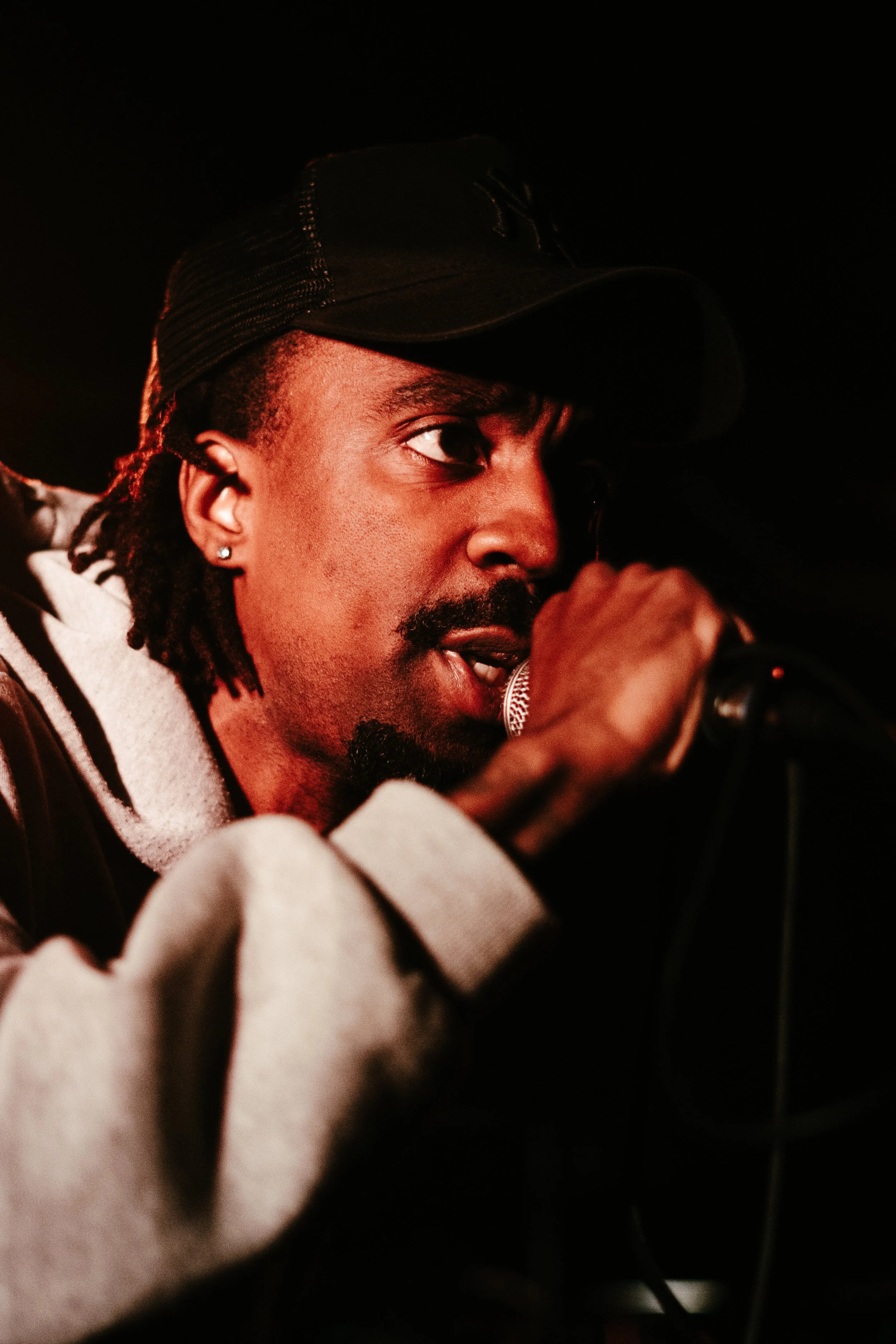 Un homme noir portant une casquette noire, chantant ou parlant dans un microphone dans un cadre sombre.