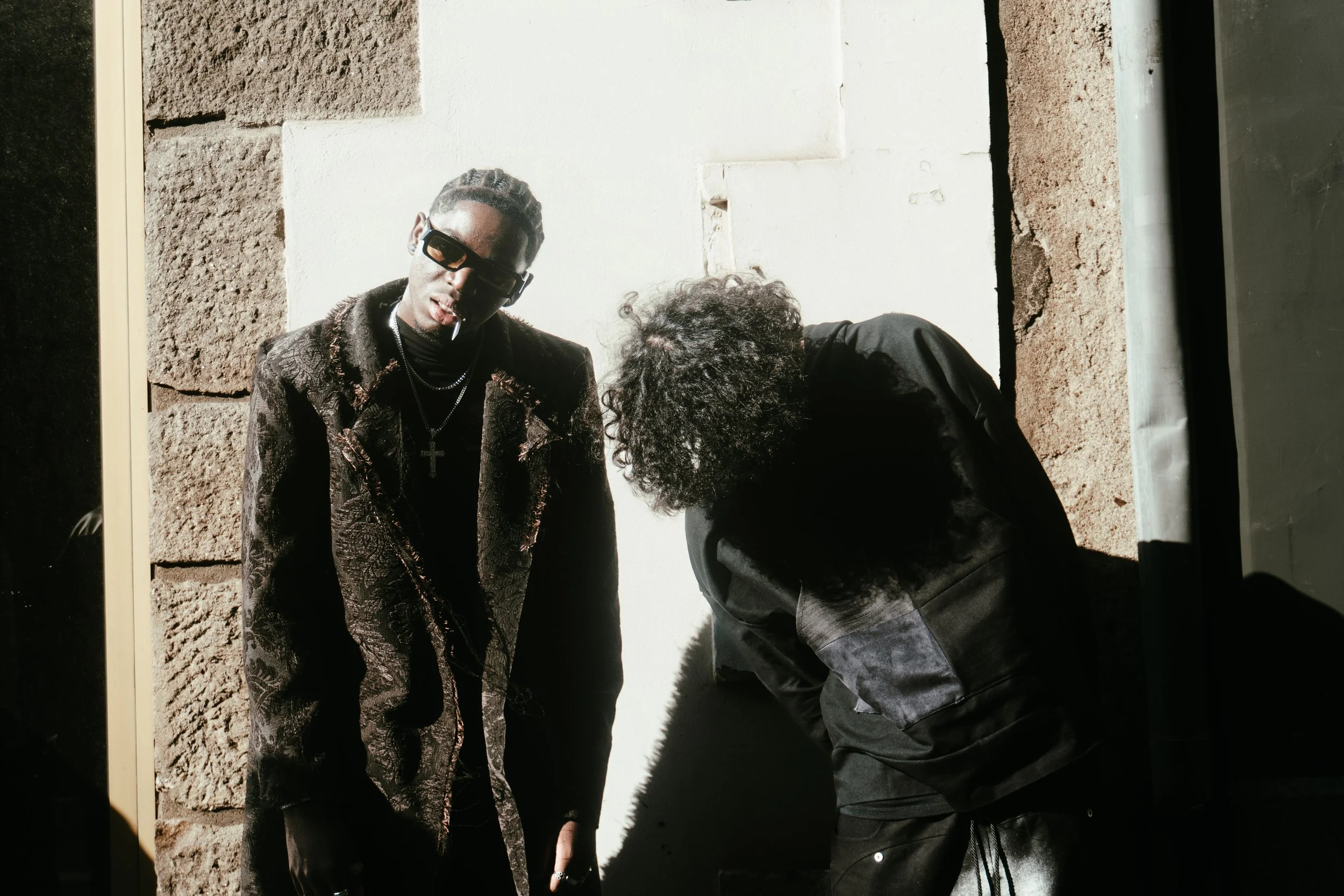 Deux hommes dans une rue, l'un porte des lunettes de soleil, un collier avec une croix, et une cigarette à la bouche, l'autre a des cheveux bouclés et porte une veste noire, ils se tiennent près d'un mur de pierre et d'un mur blanc.