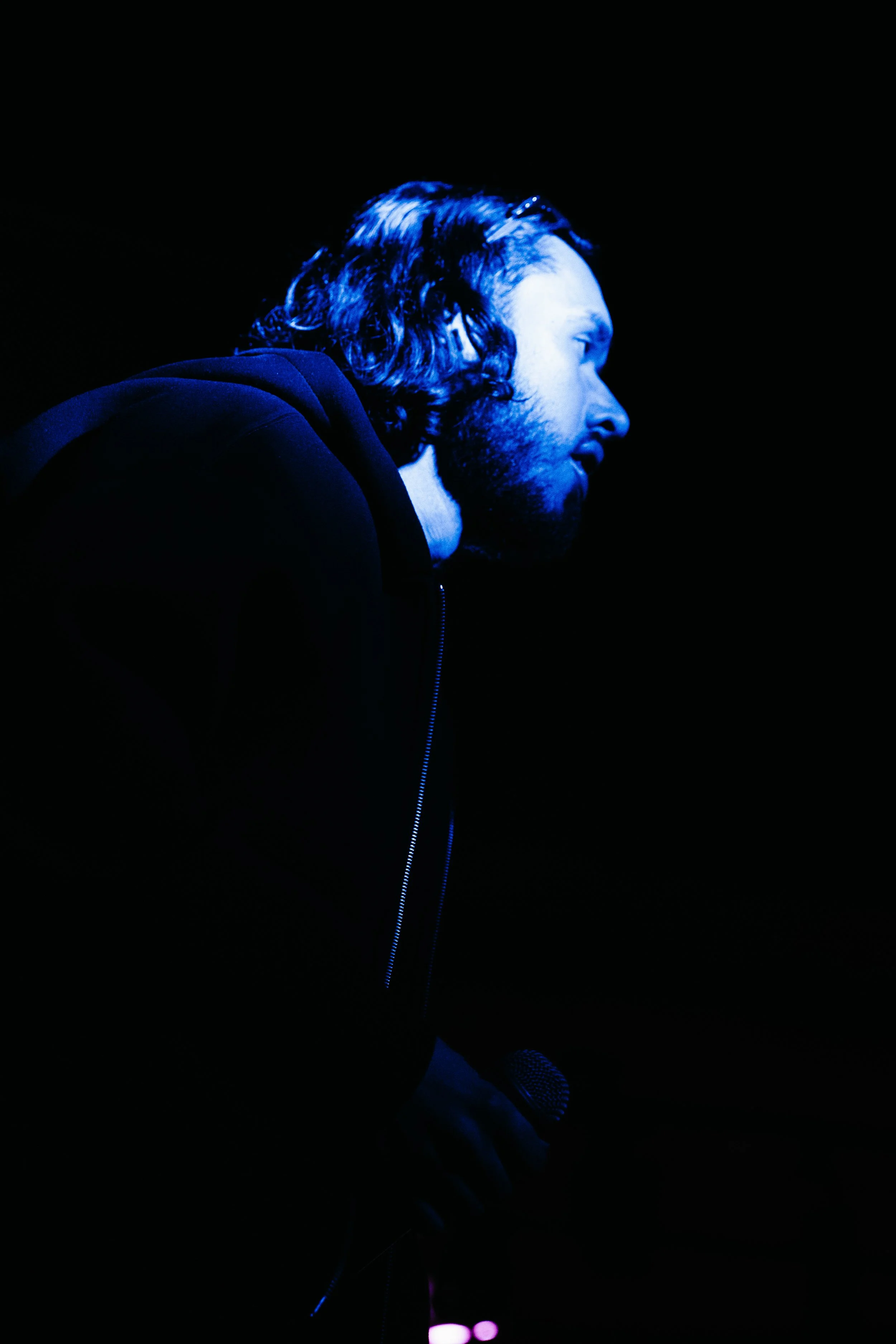 Un homme blond avec barbe, portant un sweat noir, tient un micro dans la main et est éclairé par une lumière bleue dans l'obscurité.