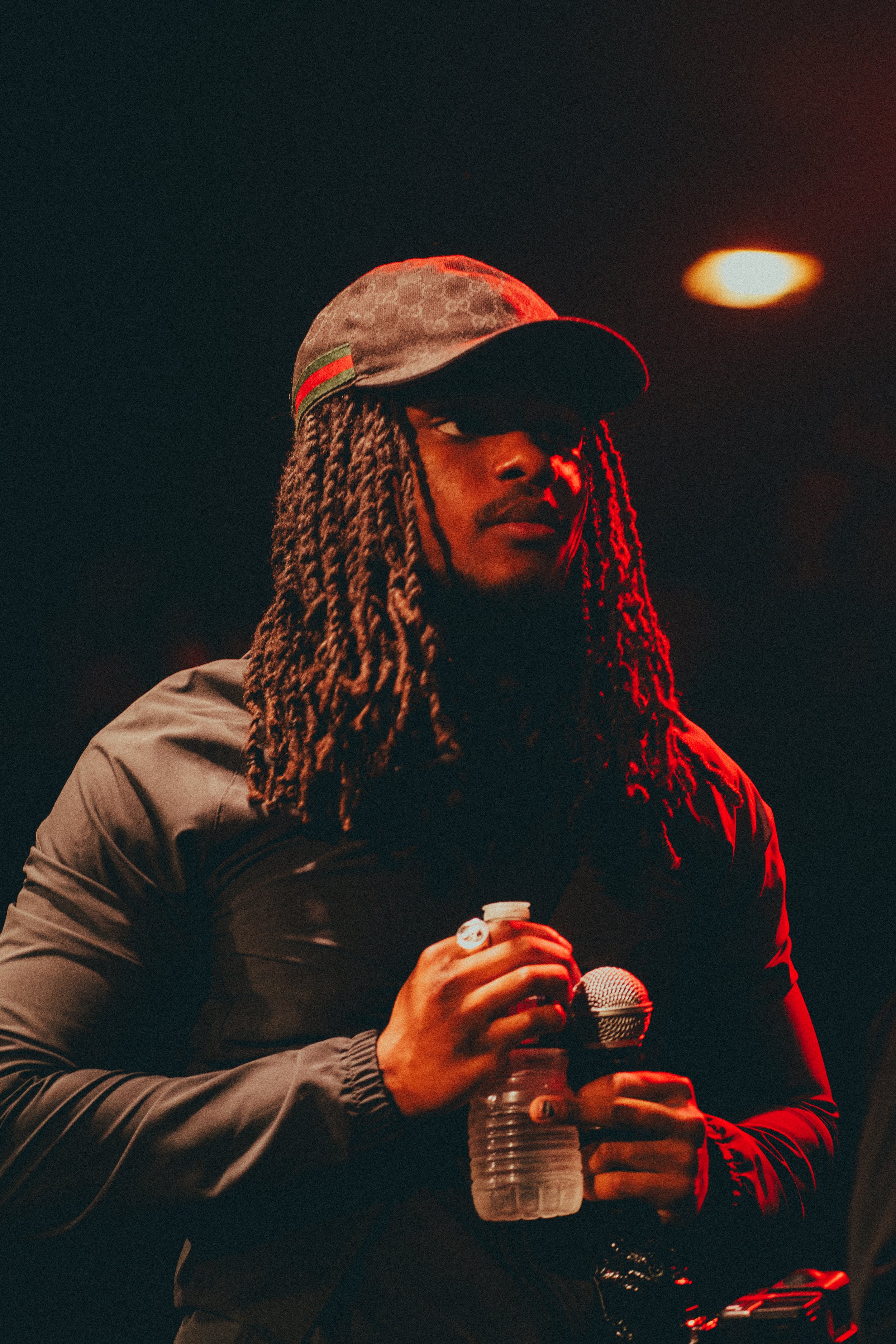 Homme avec dreadlocks portant un chapeau de baseball, tenant une bouteille d'eau et un microphone dans une scène sombre.