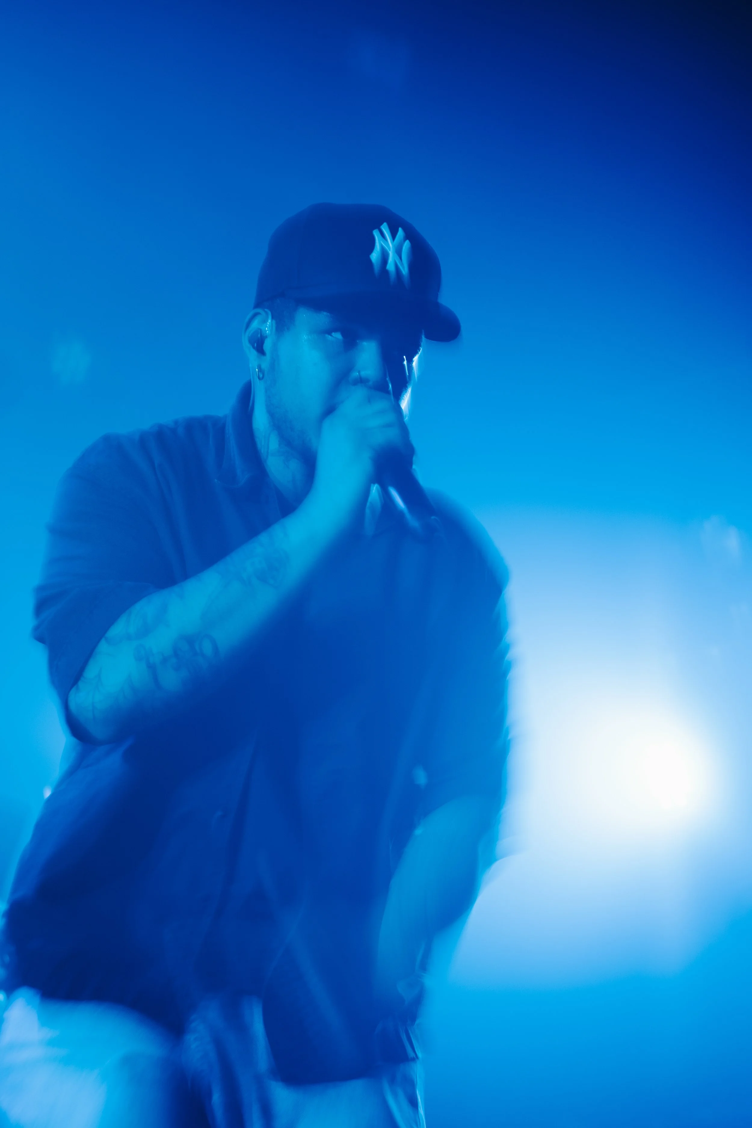 Un homme portant une casquette de baseball NYC chante ou parle dans un micro, entouré d'éclairage bleu, lors d'un concert.