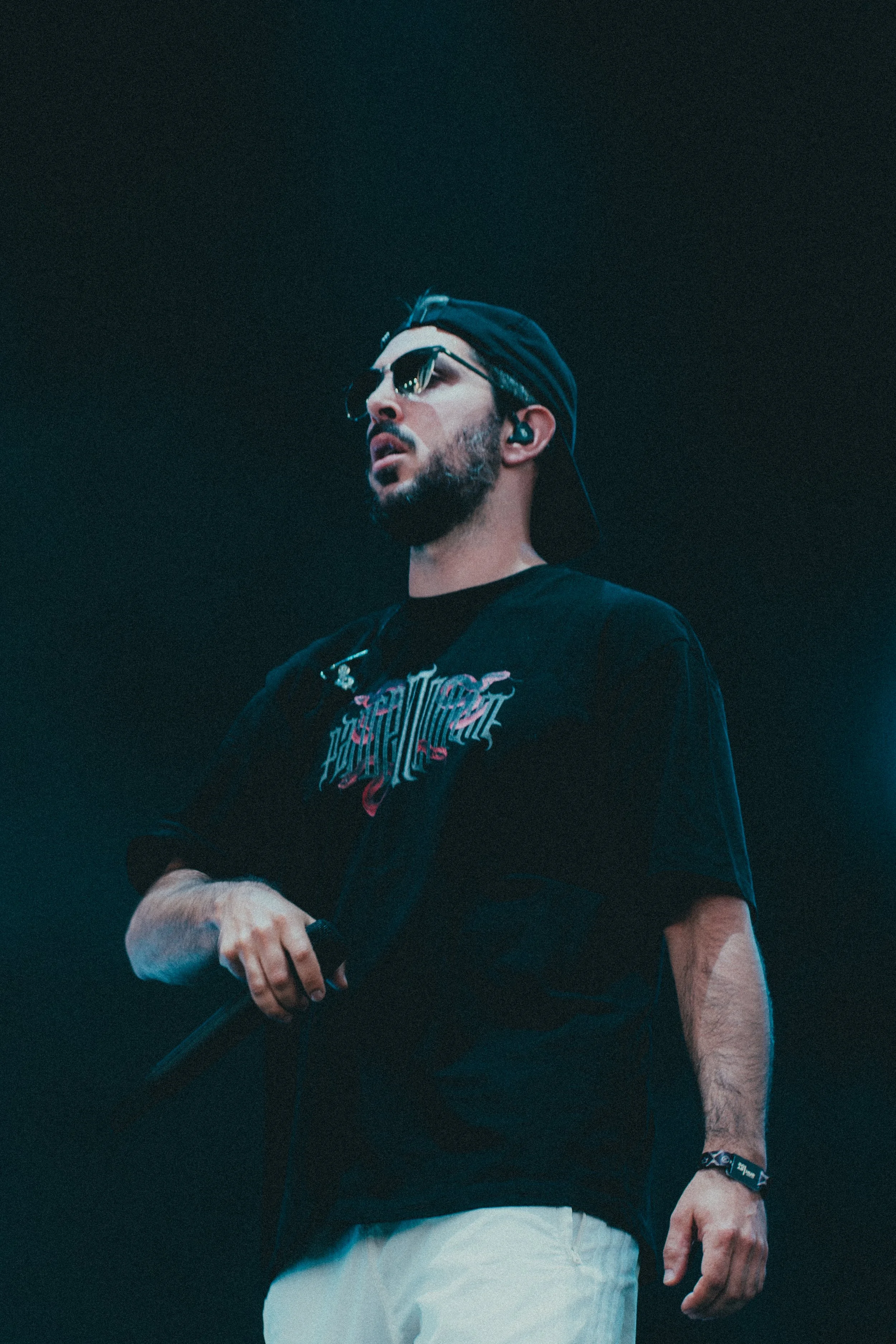Un homme avec une barbe et des lunettes de soleil, portant une casquette à l'envers, vêtue d'un t-shirt noir avec un design graphique, tient un micro dans la main, lors d'un concert ou d'une performance.