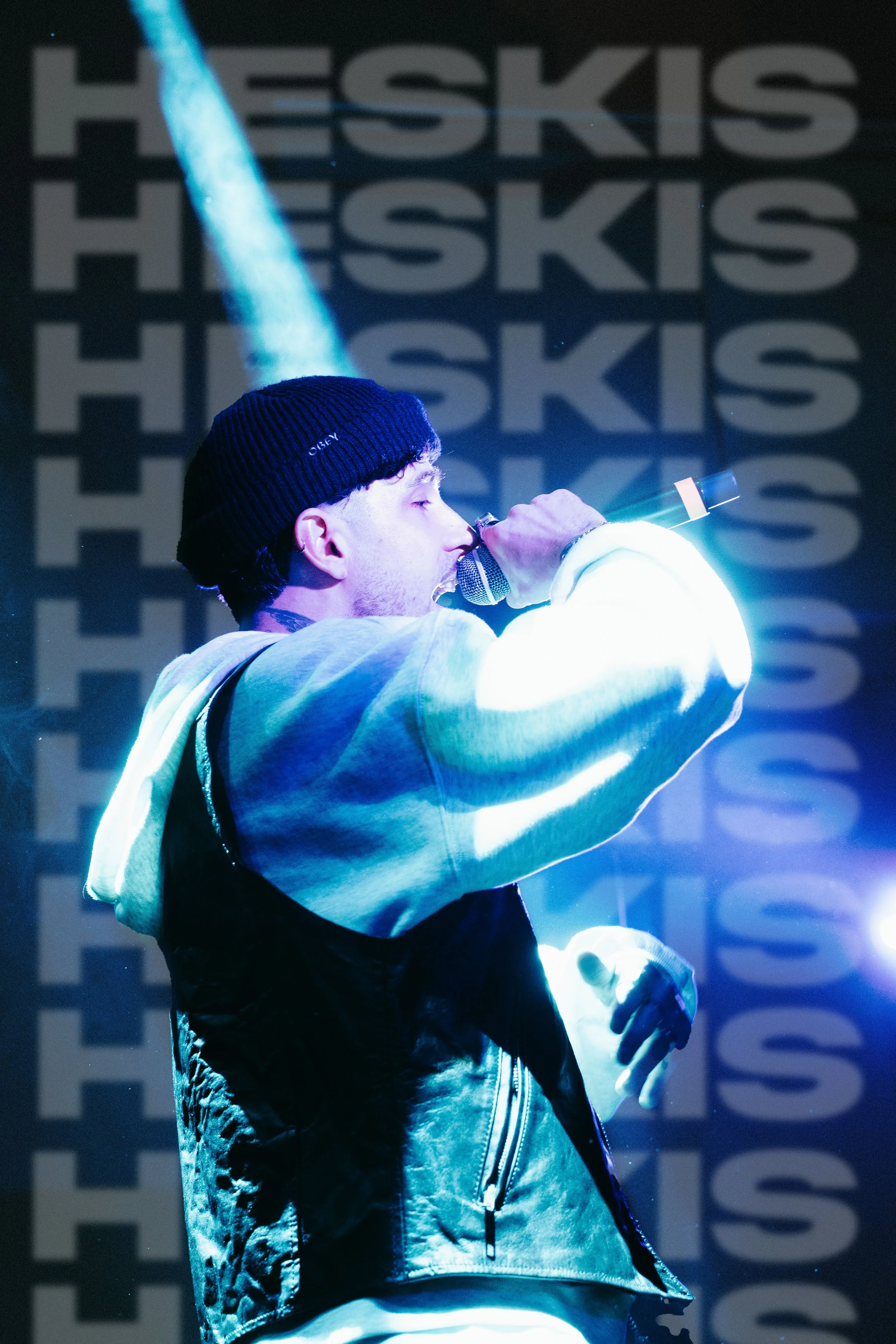 Un artiste performe sur scène, tenant un microphone, avec un fond affichant plusieurs fois le nom 'HESKIS' et un rayon de lumière bleu.