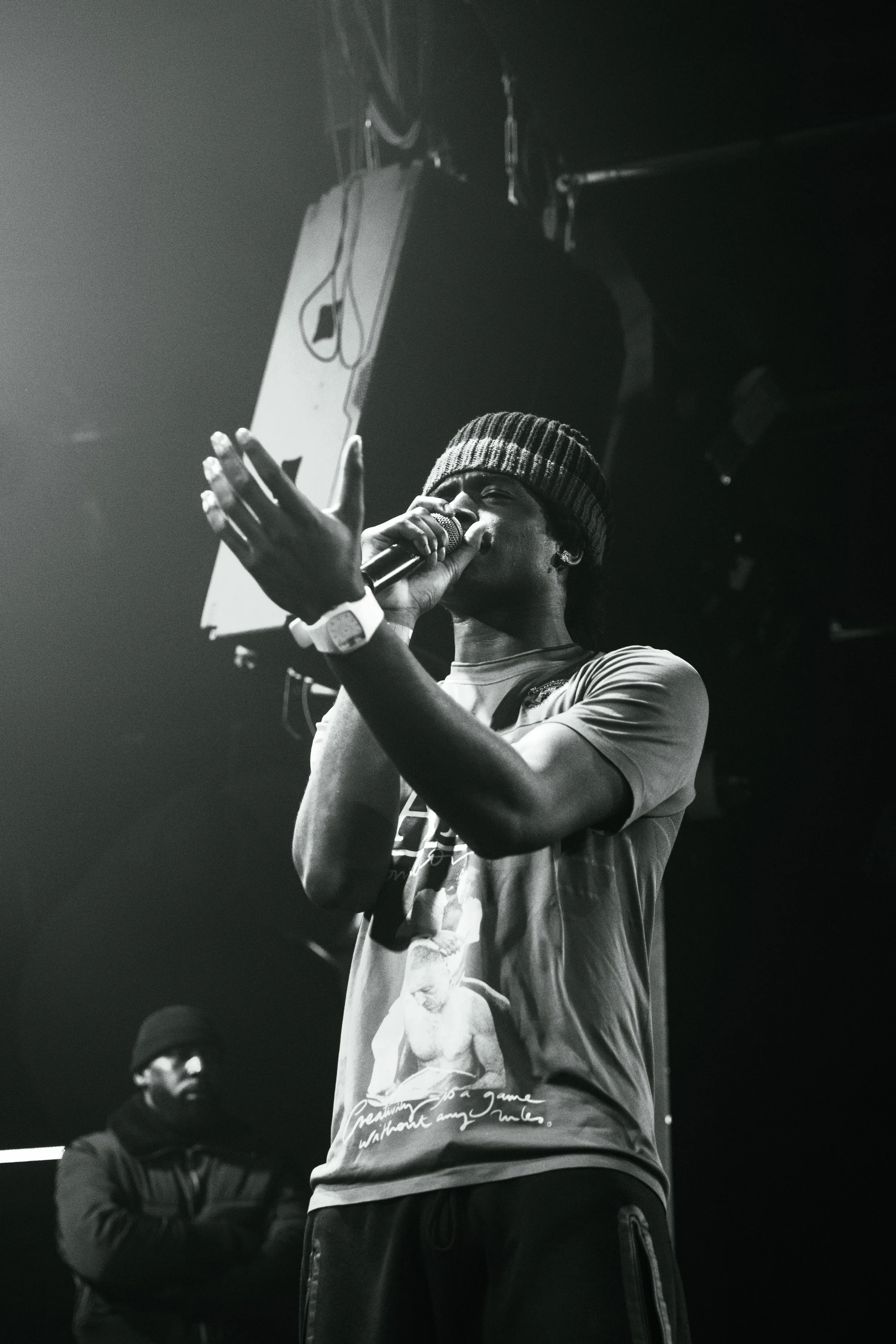 Un jeune homme portant une casquette, rapproché du microphone, semble faire un discours ou chanter sur scène, avec un autre homme en arrière-plan dans l'ombre.