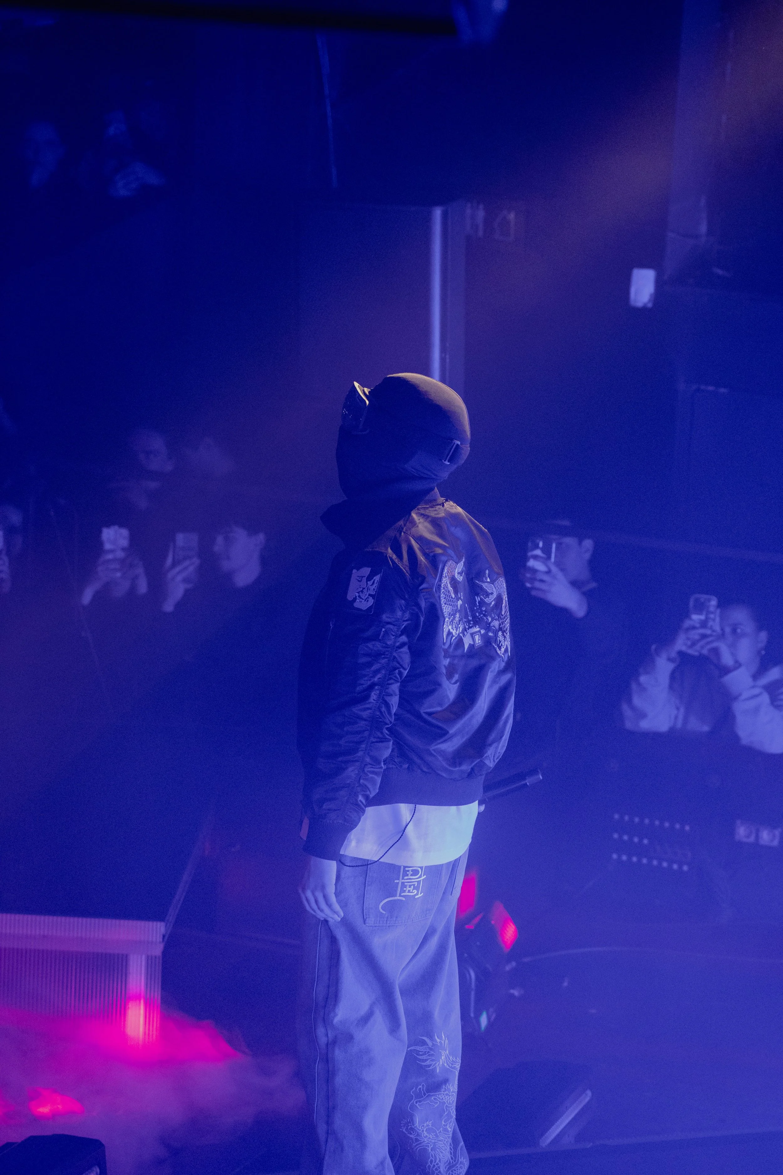 Un homme portant un casque et une veste en cuir se tient sur scène avec des vidéos de spectateurs projetées en arrière-plan, ambiance sombre et éclairage violet.