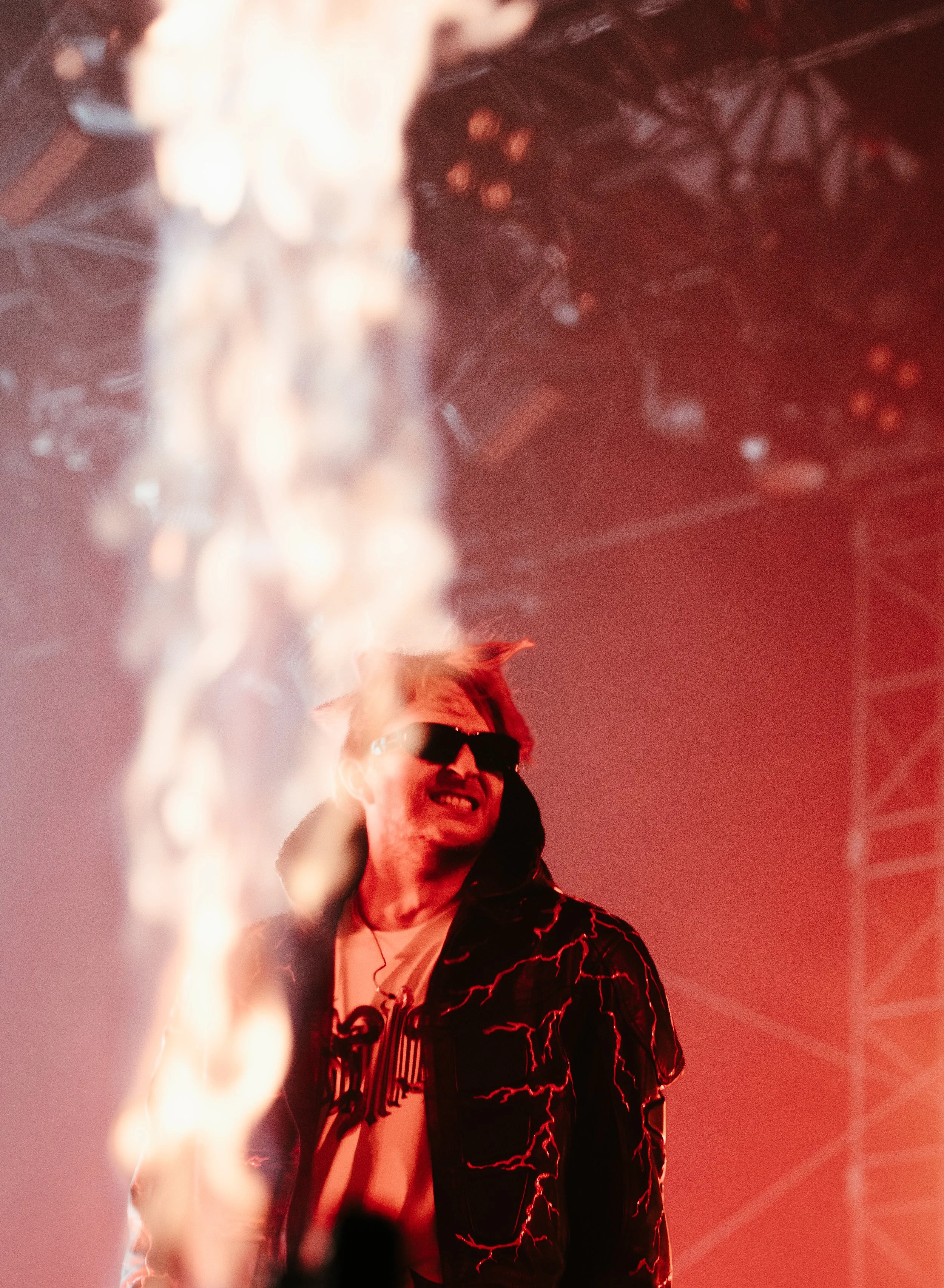 Un homme avec des cheveux blonds, portant des lunettes de soleil noires, une veste noire avec des motifs de lightning rouge, et une chemise blanche avec un logo, en train de performer sur scène avec un éclairage rouge en arrière-plan.