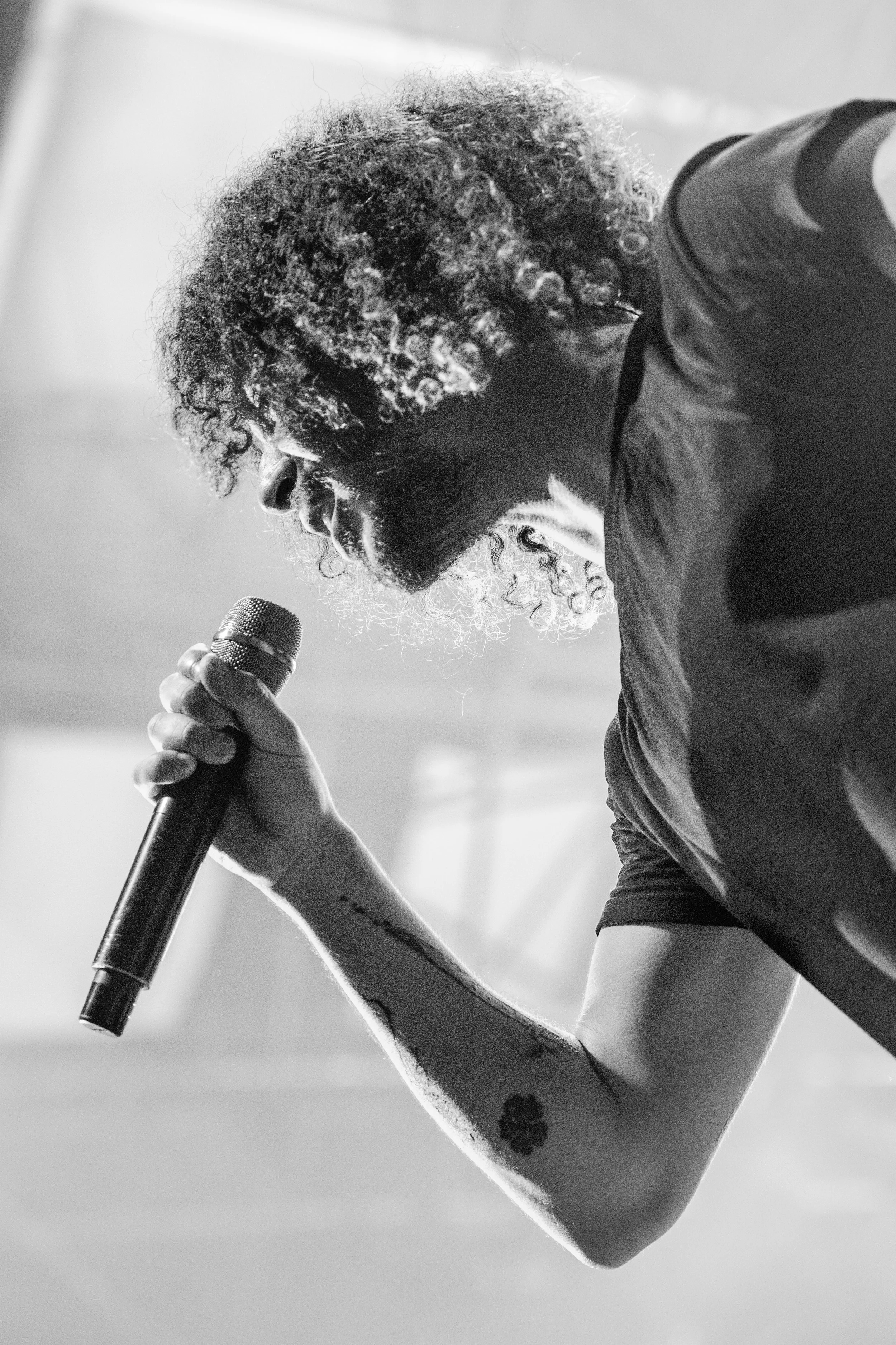Un homme avec une barbe et des cheveux bouclés, tenant un microphone, en train de chanter ou de parler, en noir et blanc.