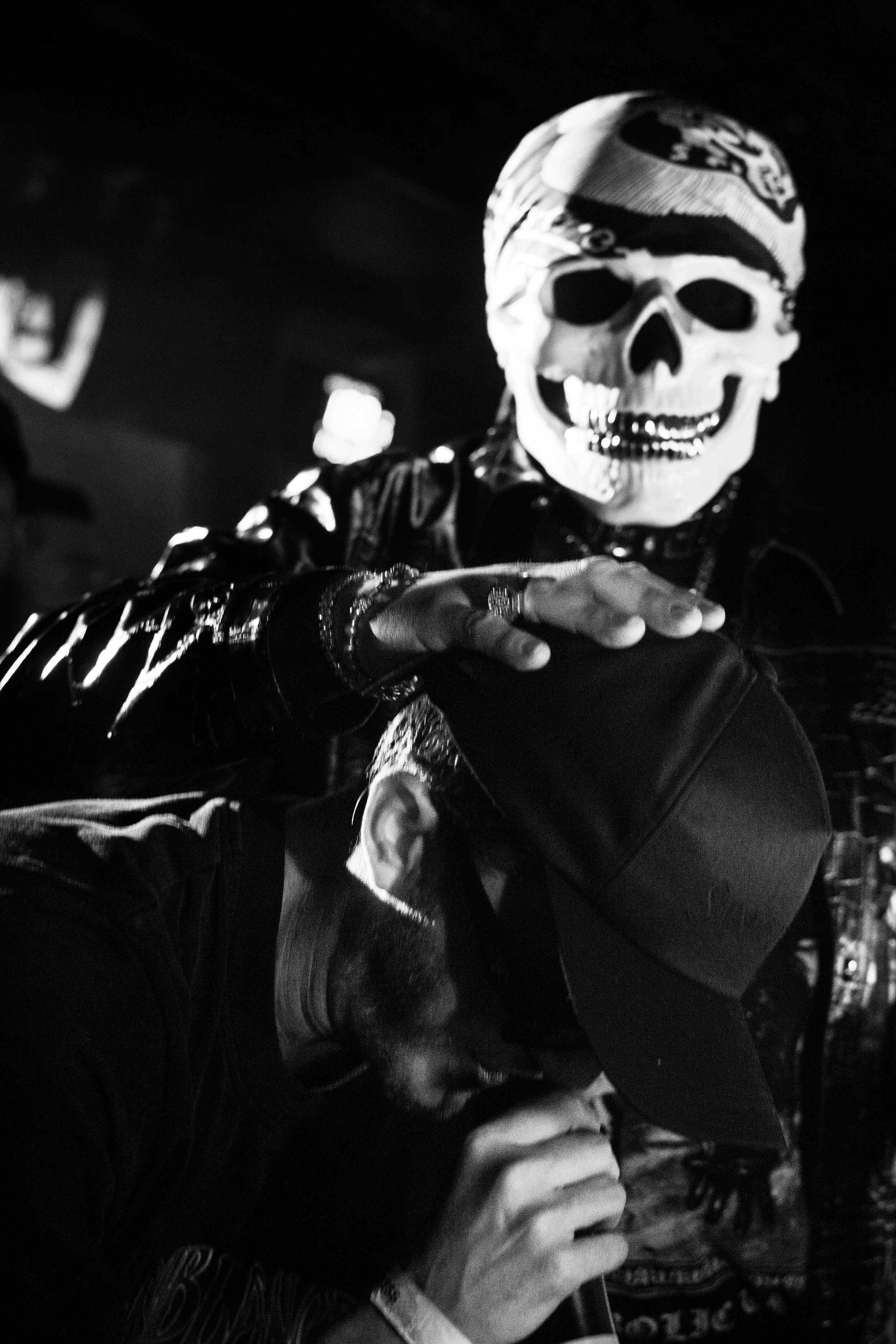 Une personne portant un masque de crâne avec bandana et un sweat à capuche noir, en train de prendre un selfie avec un autre individu portant une casquette noire dans un environnement sombre.